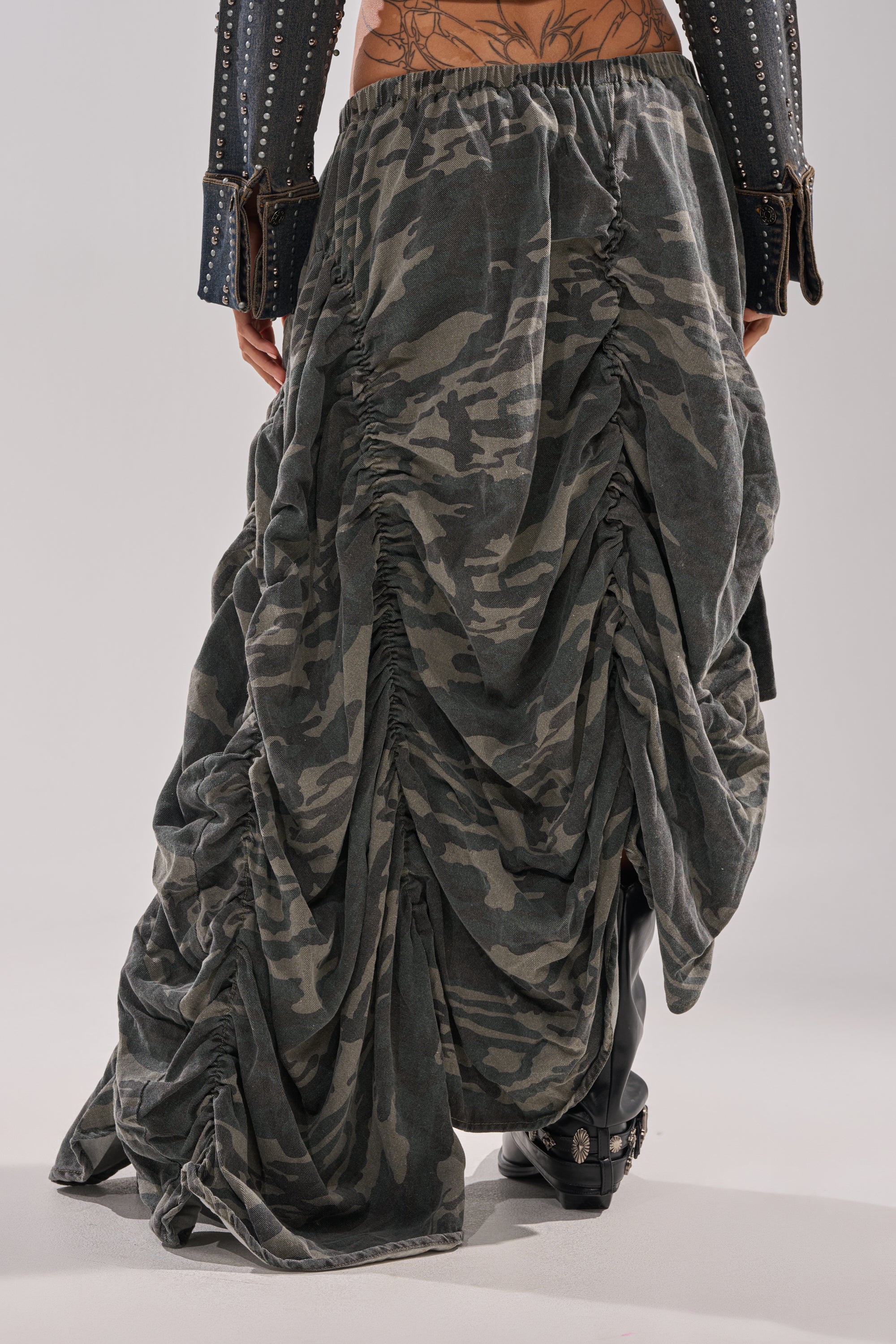 A person wearing the LETS CHANGE IT UP RUCHED MIDI SKIRT IN CAMO with black boots and a studded jacket. The photo, shot from behind, highlights the streetwear skirt’s unique ruching and a tattoo on the lower back.