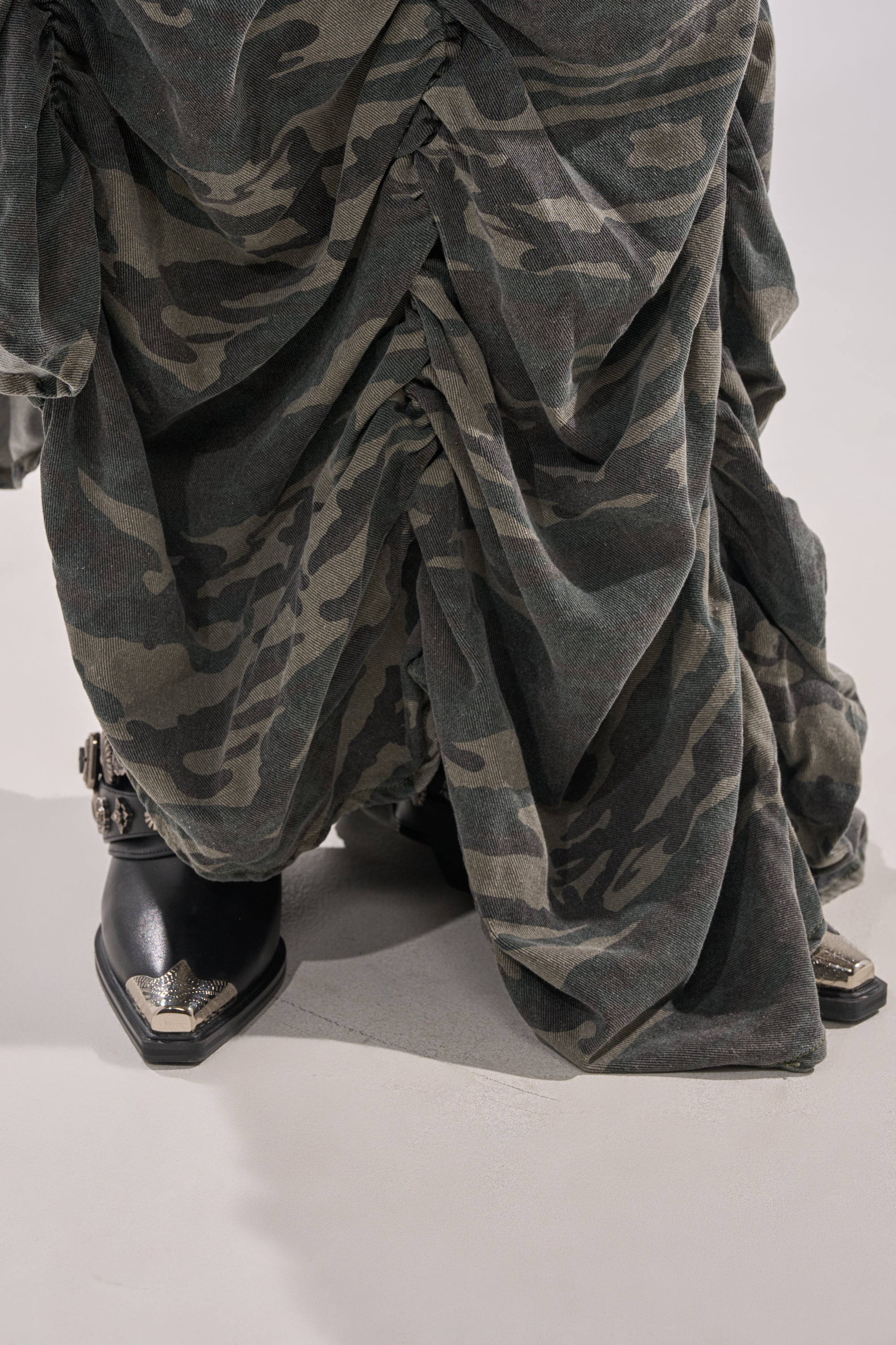 A close-up of the LETS CHANGE IT UP RUCHED MIDI SKIRT IN CAMO draped over legs, paired with black pointed-toe boots with silver toe and buckle details—a bold streetwear statement.