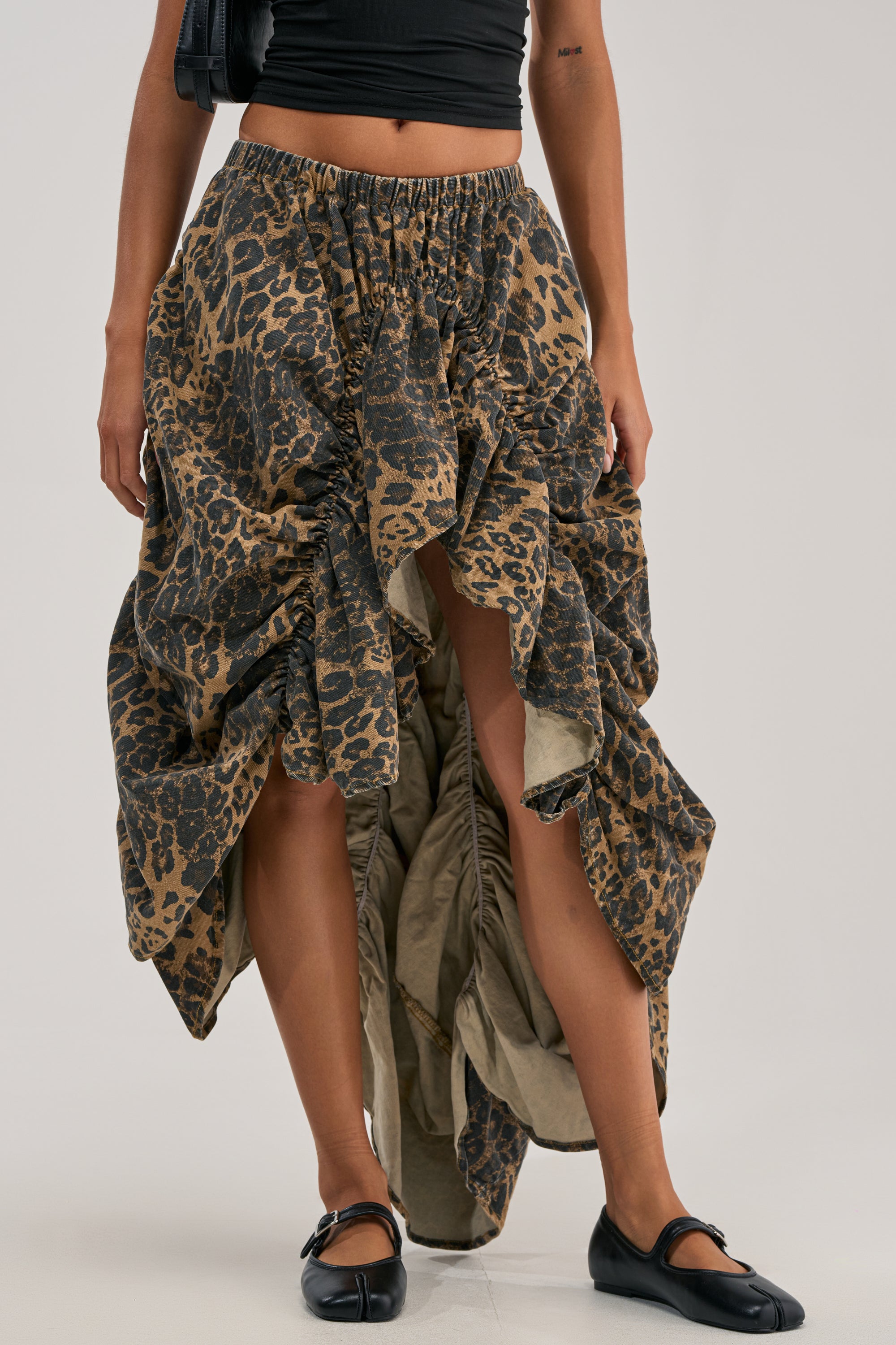 A person wears the IN TO THE WILD CHEETAH RUCHED MIDI SKIRT styled with a black crop top and black ballet flats, creating an edgy festival streetwear look against a simple background.