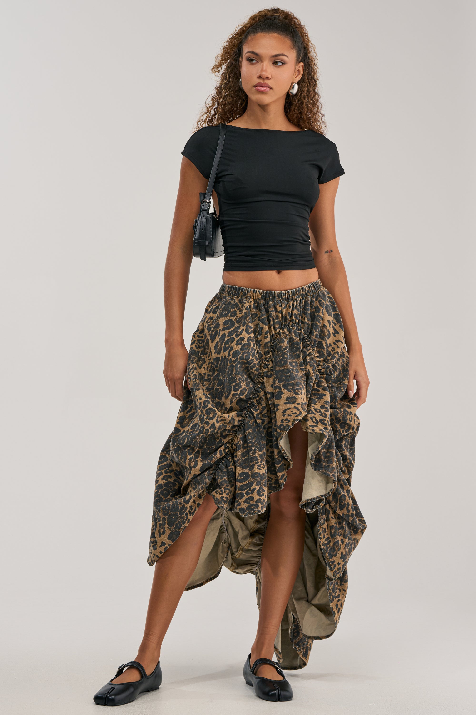 Woman in a black fitted top and the IN TO THE WILD CHEETAH RUCHED MIDI SKIRT, paired with black flats and a small black shoulder bag, standing against a plain background.