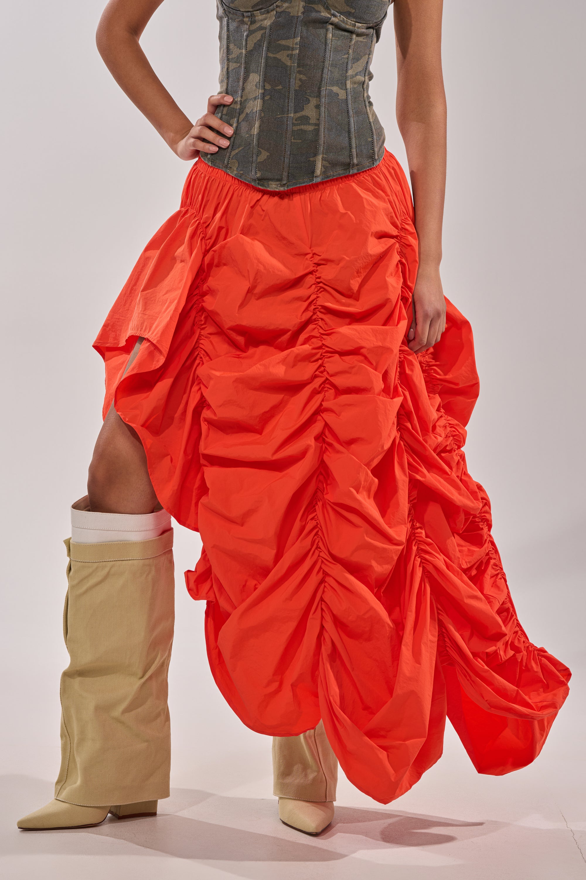 Wearing the LETS CHANGE IT UP RUCHED MIDI SKIRT IN ORANGE with a blue denim corset and oversized beige pointed-toe boots, a person poses against a plain background with their face not shown.