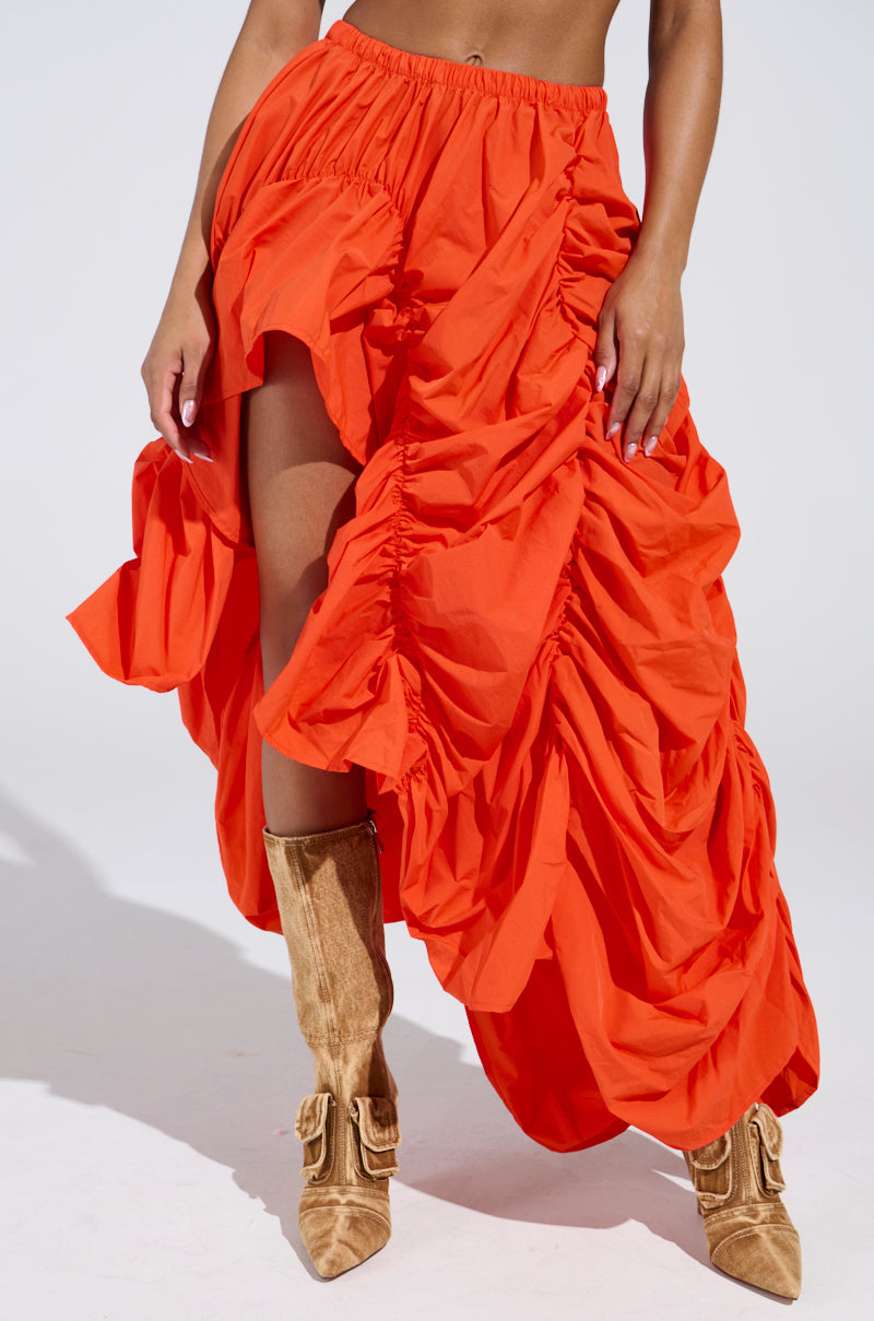 A person dons a LETS CHANGE IT UP ruched midi skirt in bright orange, featuring a high slit, paired with brown suede pointed-toe boots against a plain white background.