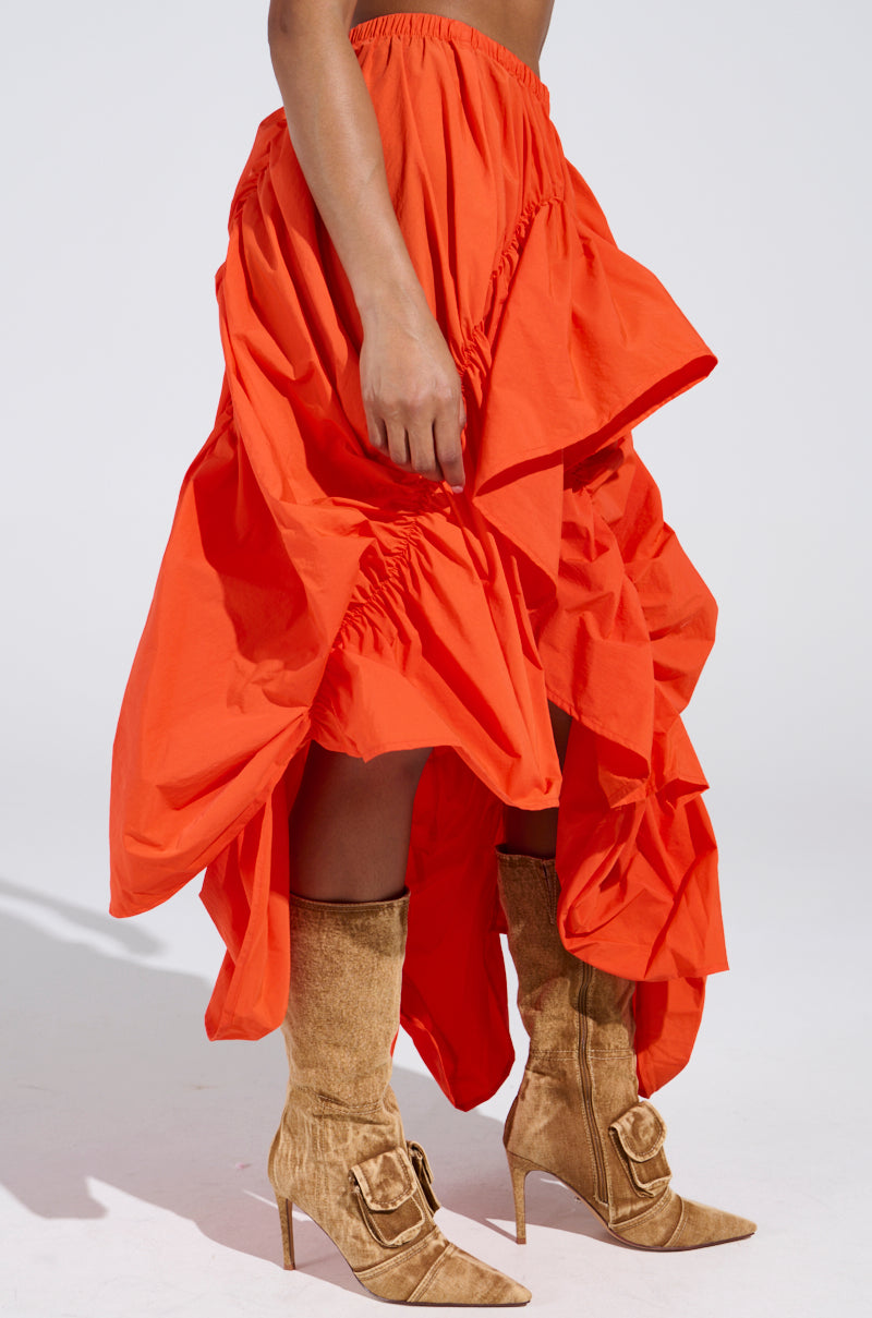 A person is wearing the LETS CHANGE IT UP RUCHED MIDI SKIRT in vibrant orange, featuring a high-low silhouette with ruffled details, and tan suede knee-high boots with decorative buckles. The background is plain white.