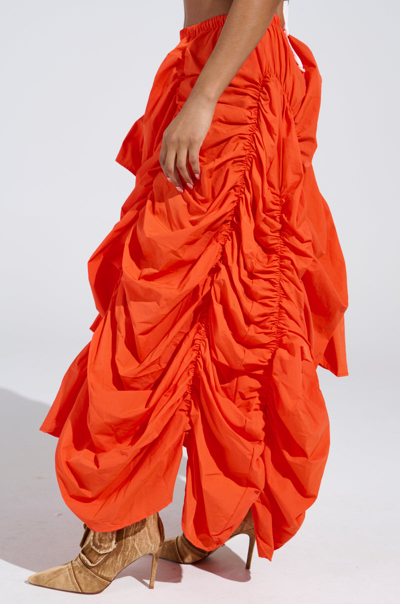 Wearing the LETS CHANGE IT UP RUCHED MIDI SKIRT IN ORANGE, featuring a vibrant hue with dramatic ruching and layered details, paired with tan suede ankle boots. Its voluminous, textured look is enhanced by a high-low silhouette against a plain white backdrop.