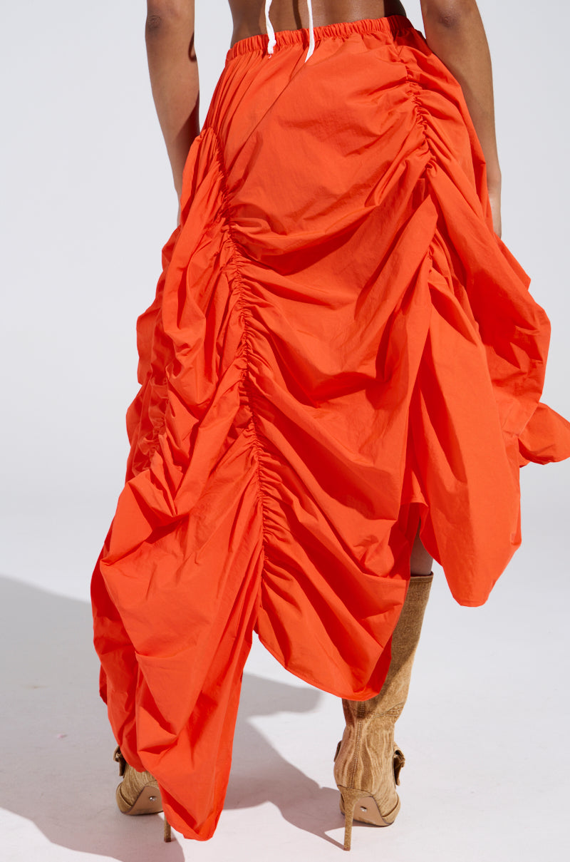 Someone wears the LETS CHANGE IT UP RUCHED MIDI SKIRT IN ORANGE, featuring a dynamic draped design with side gathers for voluminous textures and a hint of high-low silhouette. It's paired perfectly with knee-high brown high-heeled boots against a plain, light background.