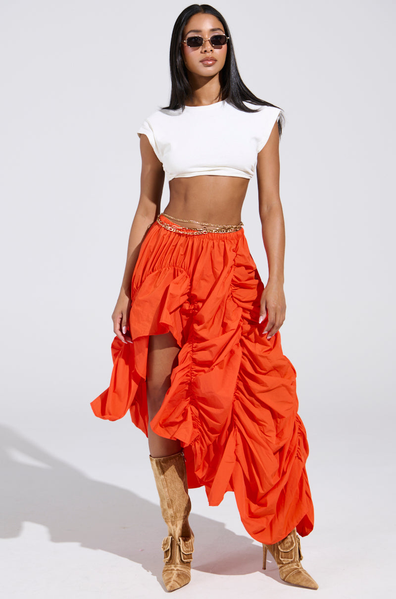 A woman confidently showcases her style wearing sunglasses, a white crop top, and the LETS CHANGE IT UP RUCHED MIDI SKIRT IN ORANGE with a high slit. She pairs it with tan knee-high boots against a simple, light background.