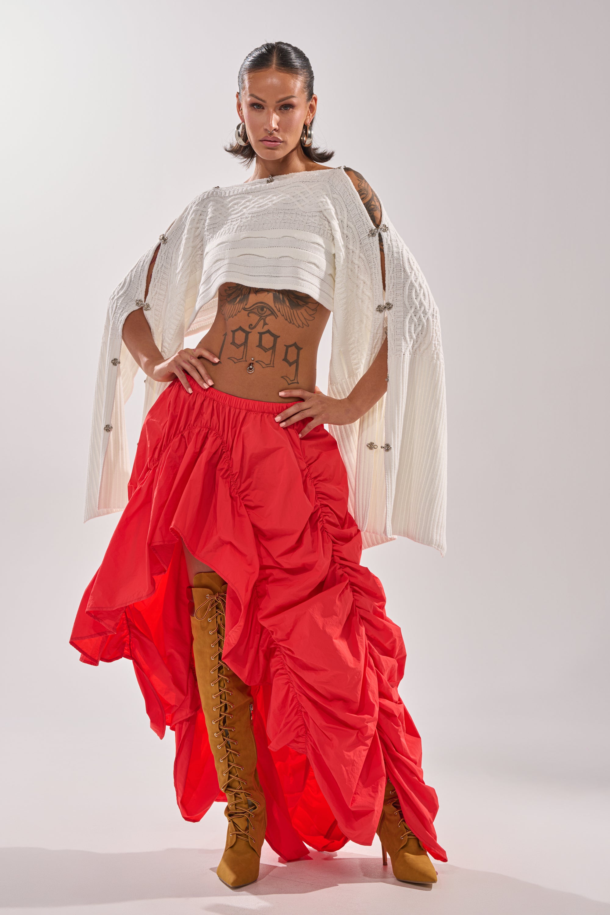 A woman stands confidently in a white textured top, tan lace-up boots, and the LETS CHANGE IT UP RUCHED MIDI SKIRT IN RED. Tattoos accent her torso and she sports short, slicked-back hair against a plain background. 0