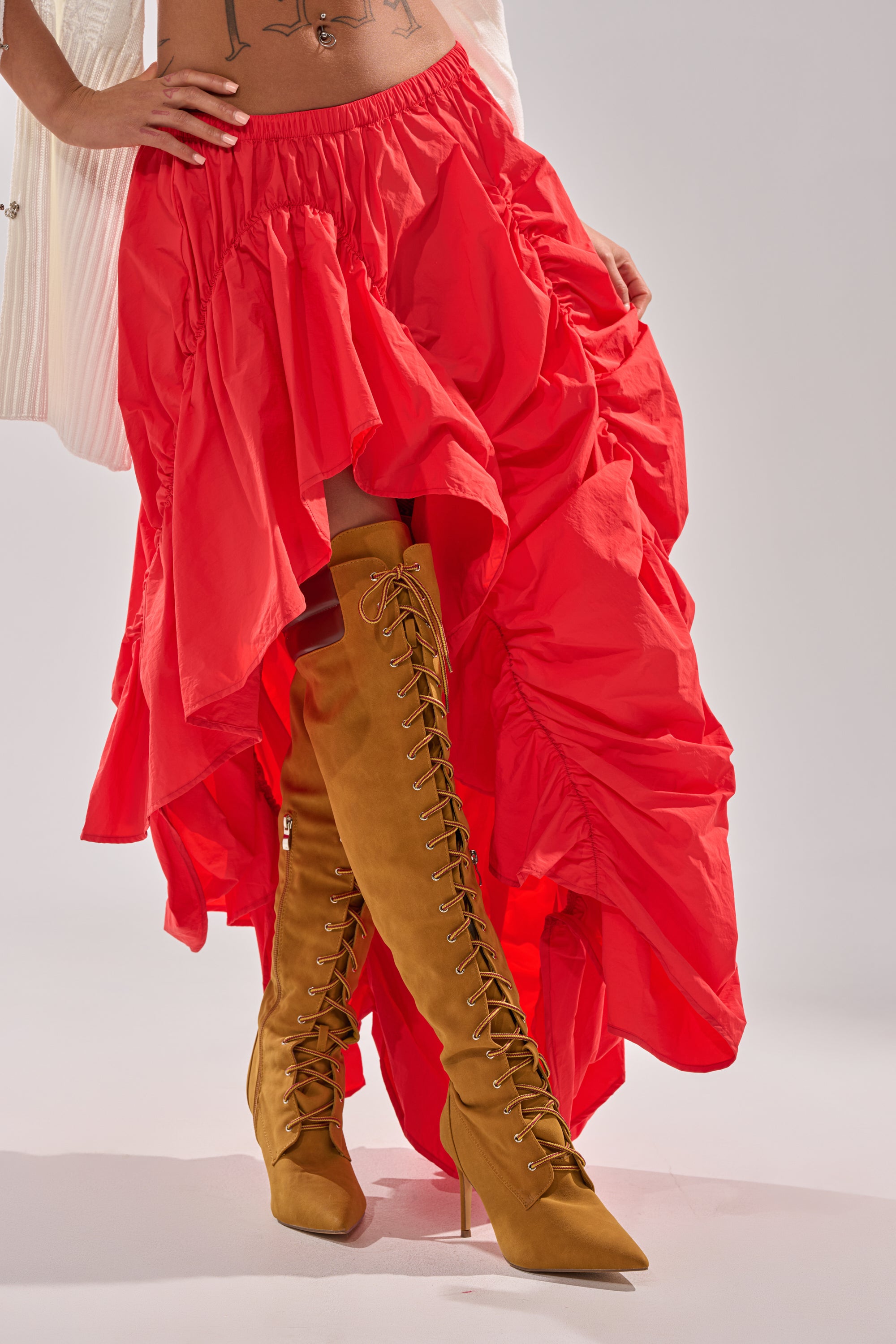 A person wears the LETS CHANGE IT UP RUCHED MIDI SKIRT IN RED with tan high-heeled lace-up boots, revealing part of their midriff and hand. The background is plain and light-colored. 2