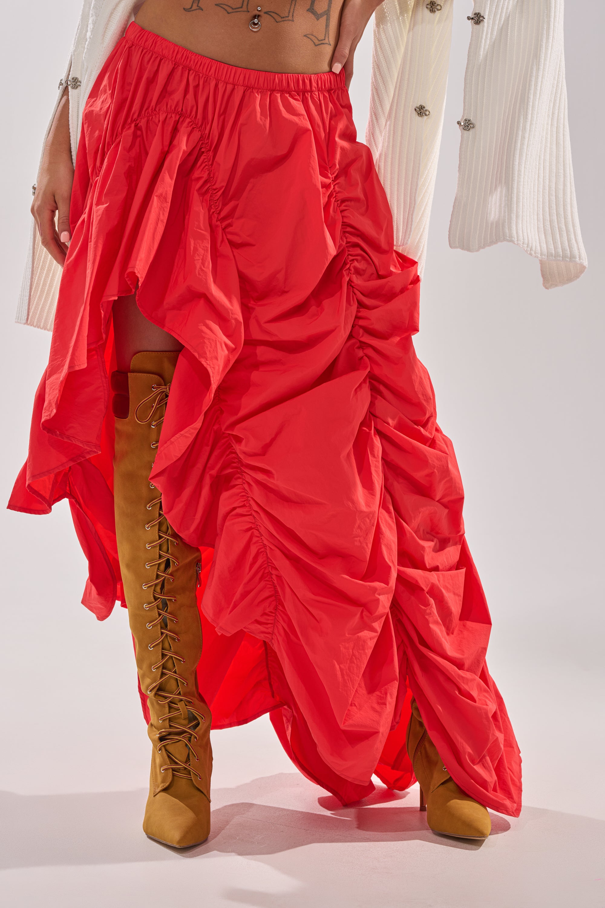A person wearing the LETS CHANGE IT UP RUCHED MIDI SKIRT IN RED, styled with knee-high tan lace-up boots. Only the lower half of the outfit is visible. 3