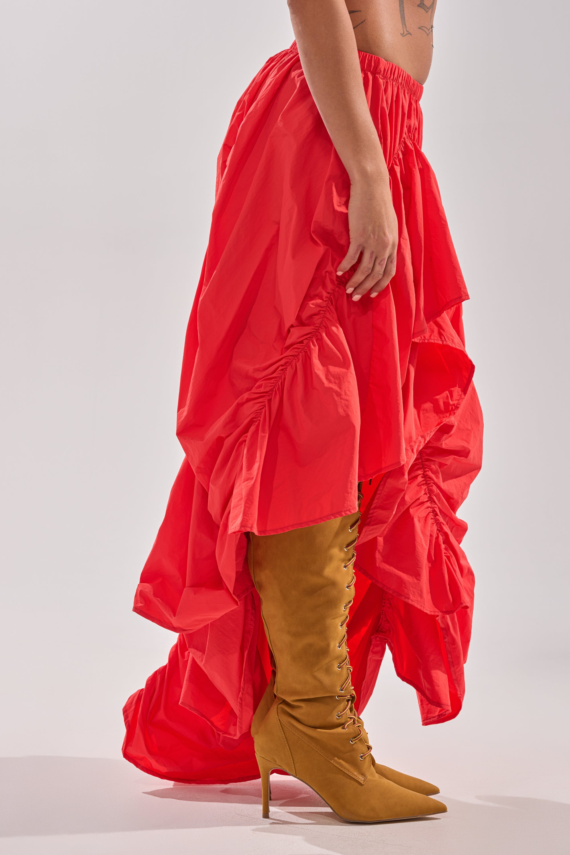 A person wearing the LETS CHANGE IT UP RUCHED MIDI SKIRT IN RED and tan knee-high, lace-up heeled boots stands against a light grey background. 4