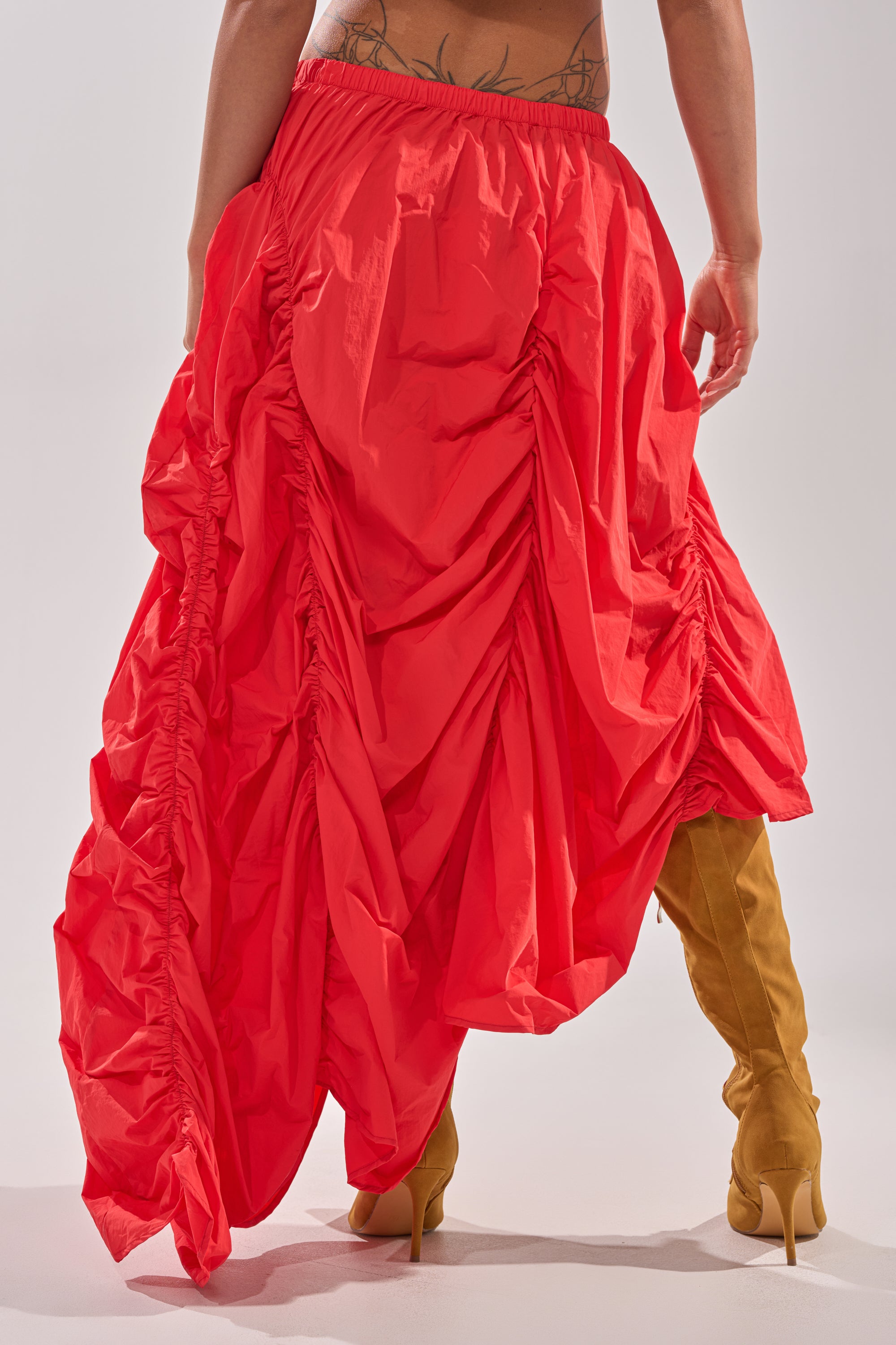 A person stands with their back to the camera, revealing lower back tattoos while wearing the LETS CHANGE IT UP RUCHED MIDI SKIRT IN RED and tan knee-high heeled boots. 5