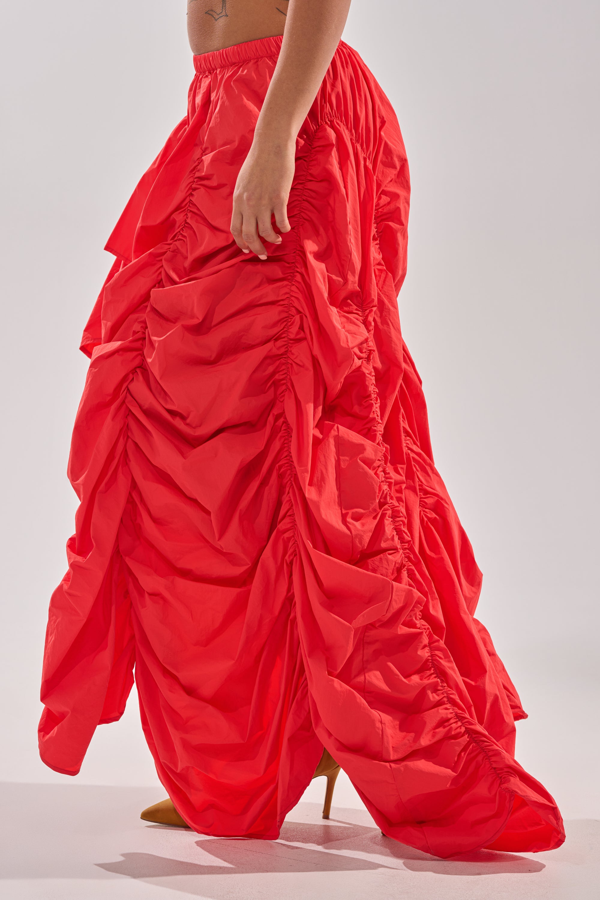 A person showcases the LETS CHANGE IT UP RUCHED MIDI SKIRT IN RED, featuring gathered details, standing in profile against a plain background with only the lower half visible. 6