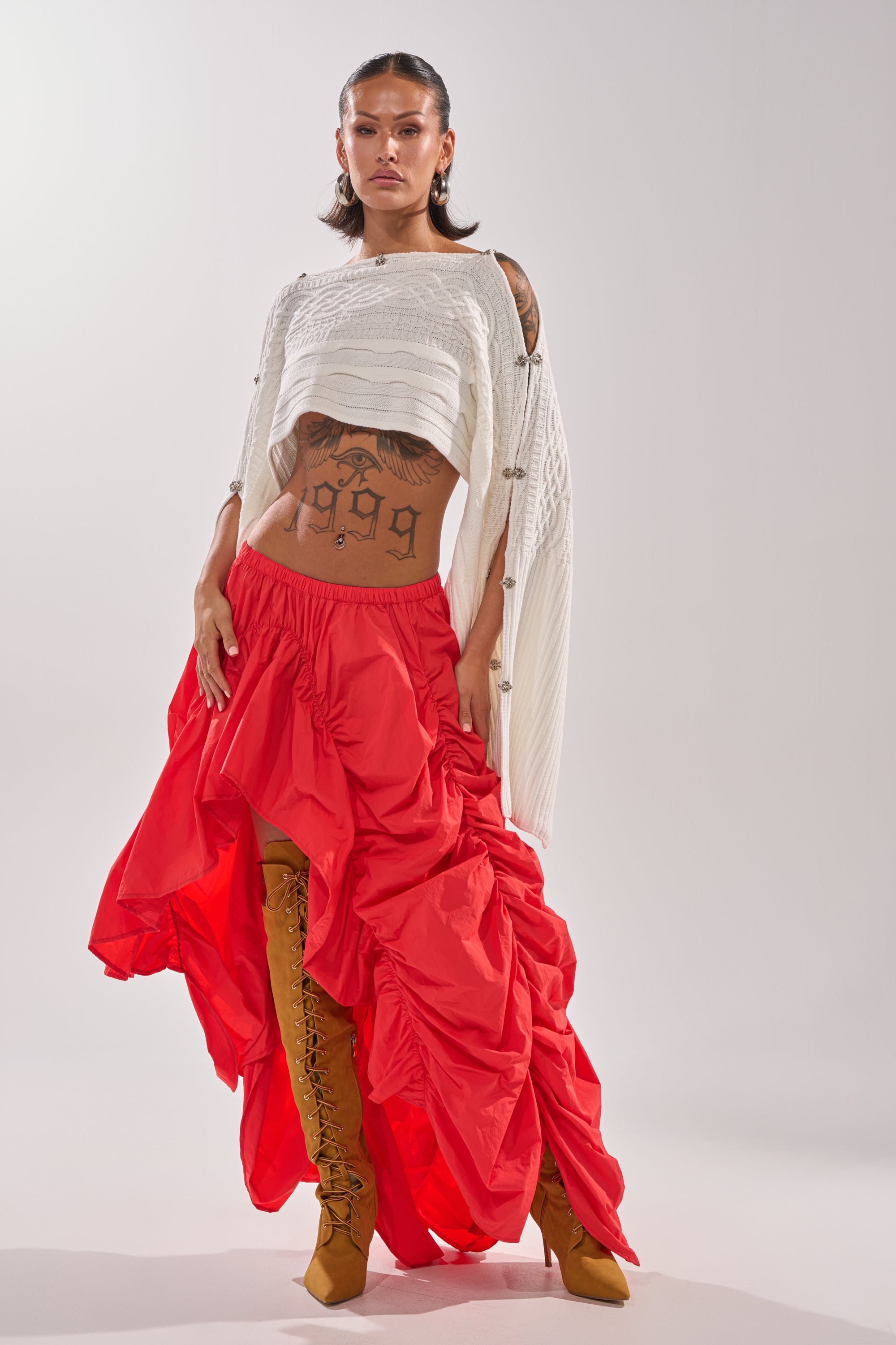 A woman stands confidently in the LETS CHANGE IT UP RUCHED MIDI SKIRT IN RED, paired with a white cropped sweater and tan lace-up boots. She has visible tattoos and slicked-back hair, set against a plain light background. 7