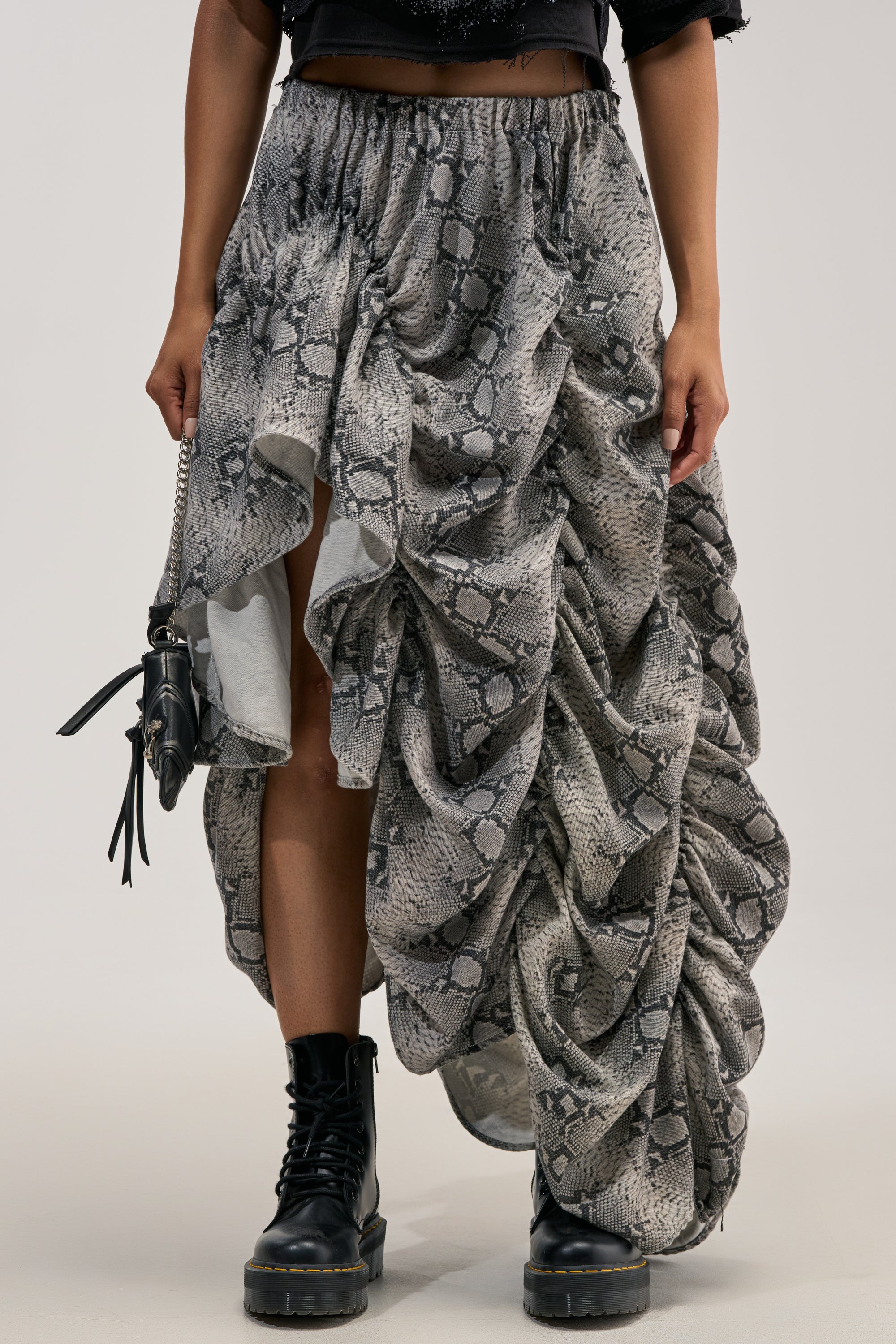 Wearing a black crop top, black combat boots, and carrying a small black bag, the person styles the LETS CHANGE IT UP RUCHED MIDI SKIRT IN SNAKE—designed with dramatic draping and a bold snakeskin texture—as the perfect festival skirt.