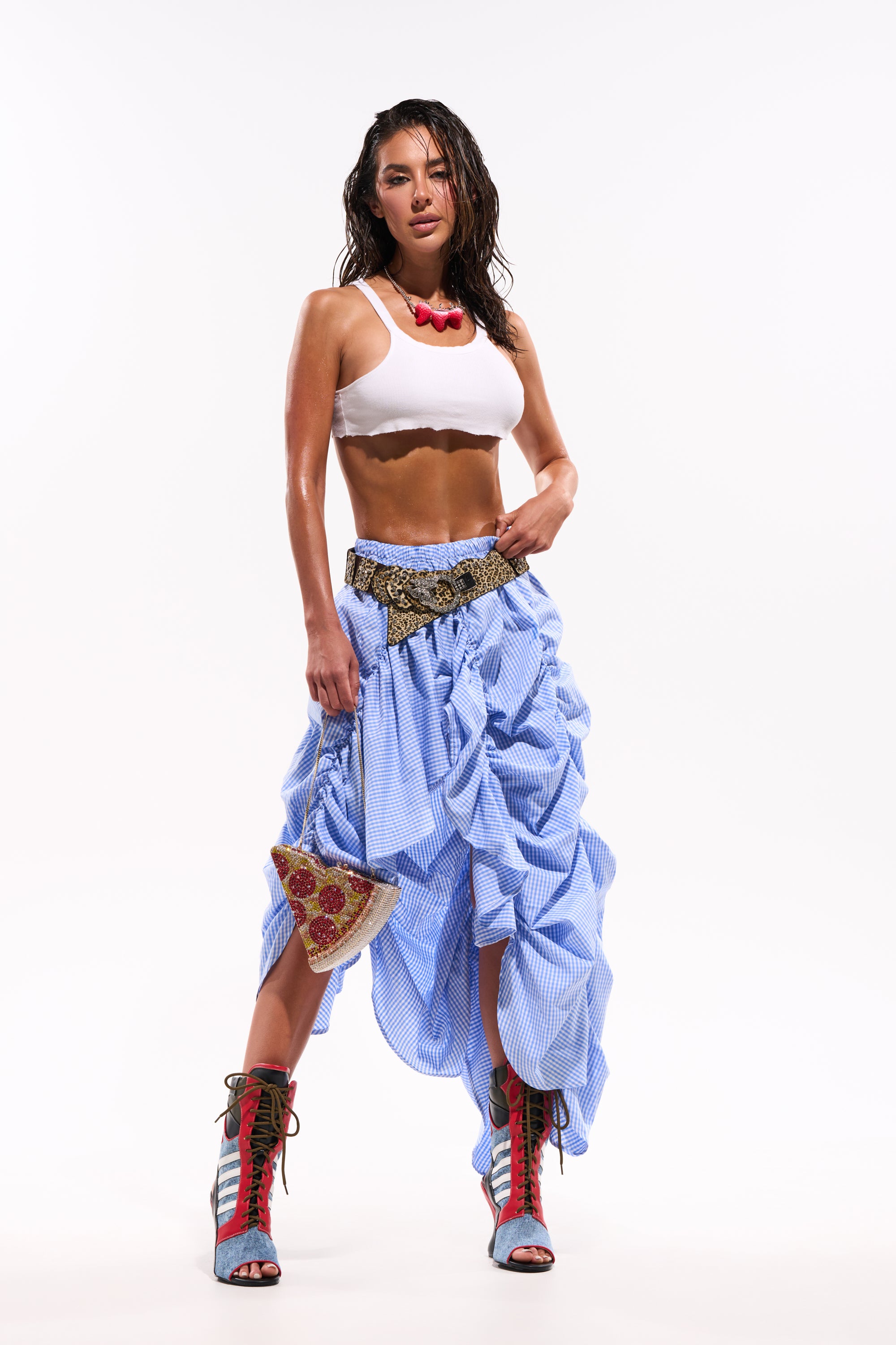 A woman stands against a white background wearing a white crop top, red choker, red and turquoise lace-up boots, holding a pizza-shaped accessory, and the COASTING STRIPE RUCHED SKIRT—a blue-and-white check skirt. 0