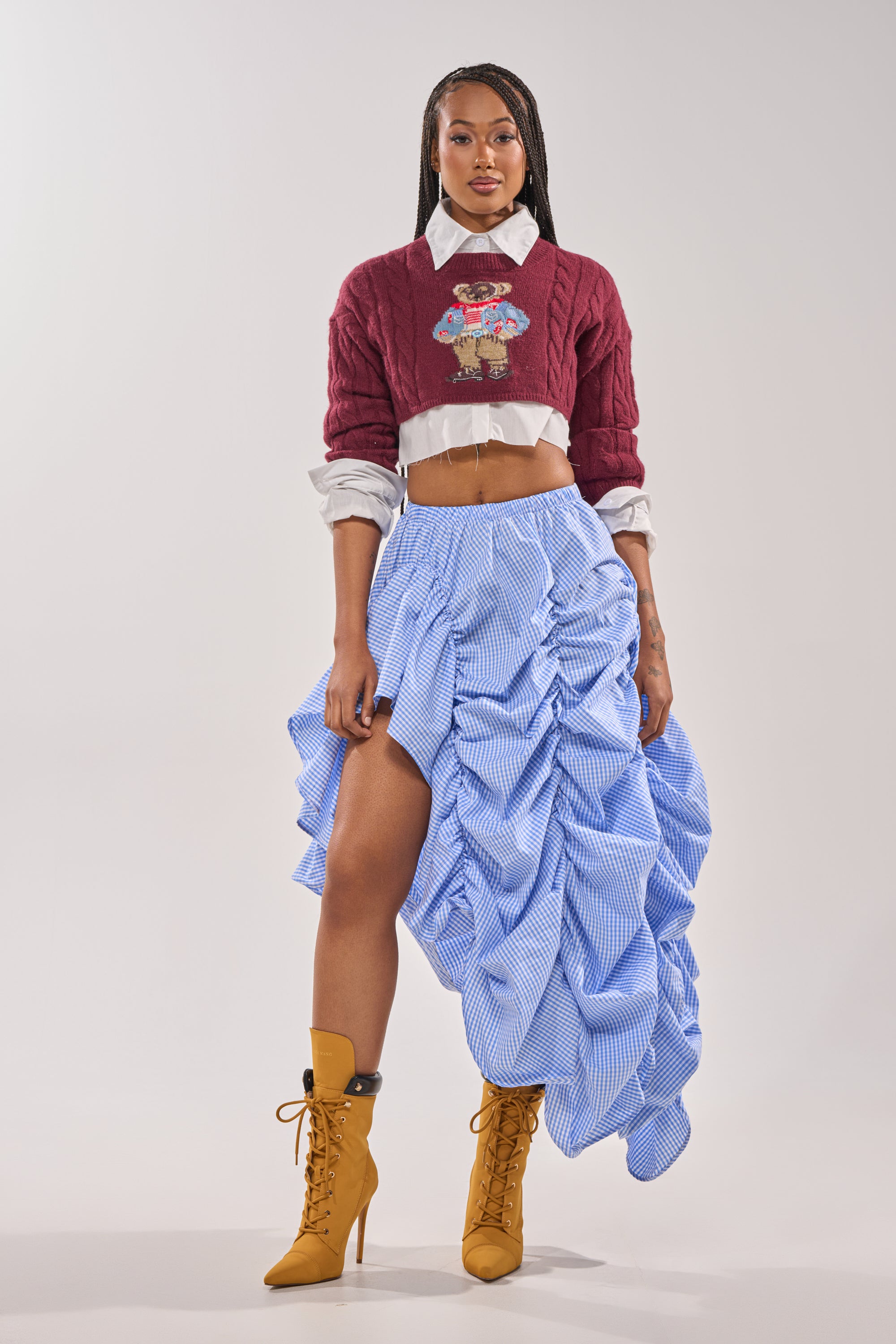 A person poses confidently in a maroon cropped sweater over a white shirt, featuring the COASTING STRIPE RUCHED SKIRT and mustard-yellow heeled boots against a plain background. 6