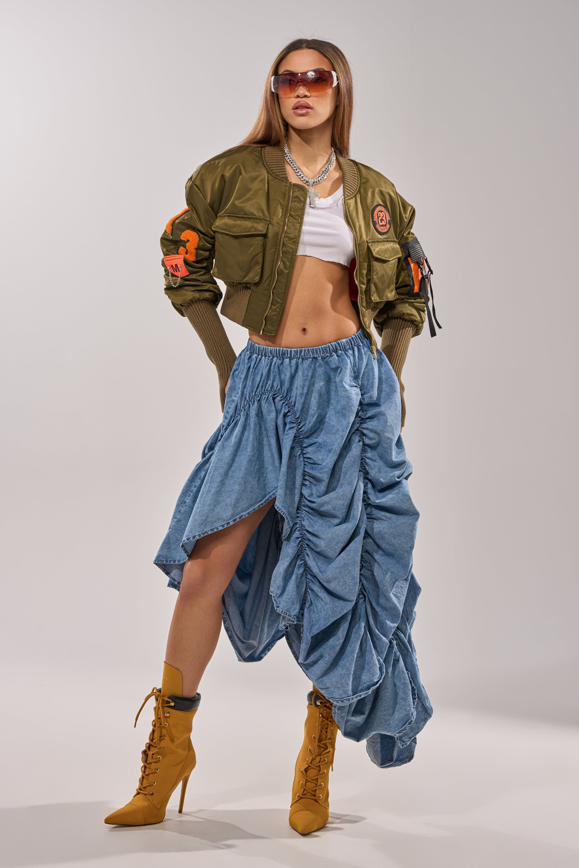 A woman poses confidently in a cropped white top, olive bomber jacket with orange details, the LETS CHANGE IT UP RUCHED MIDI SKIRT IN CHAMBRAY, mustard lace-up high-heeled boots, sunglasses, and layered necklaces.