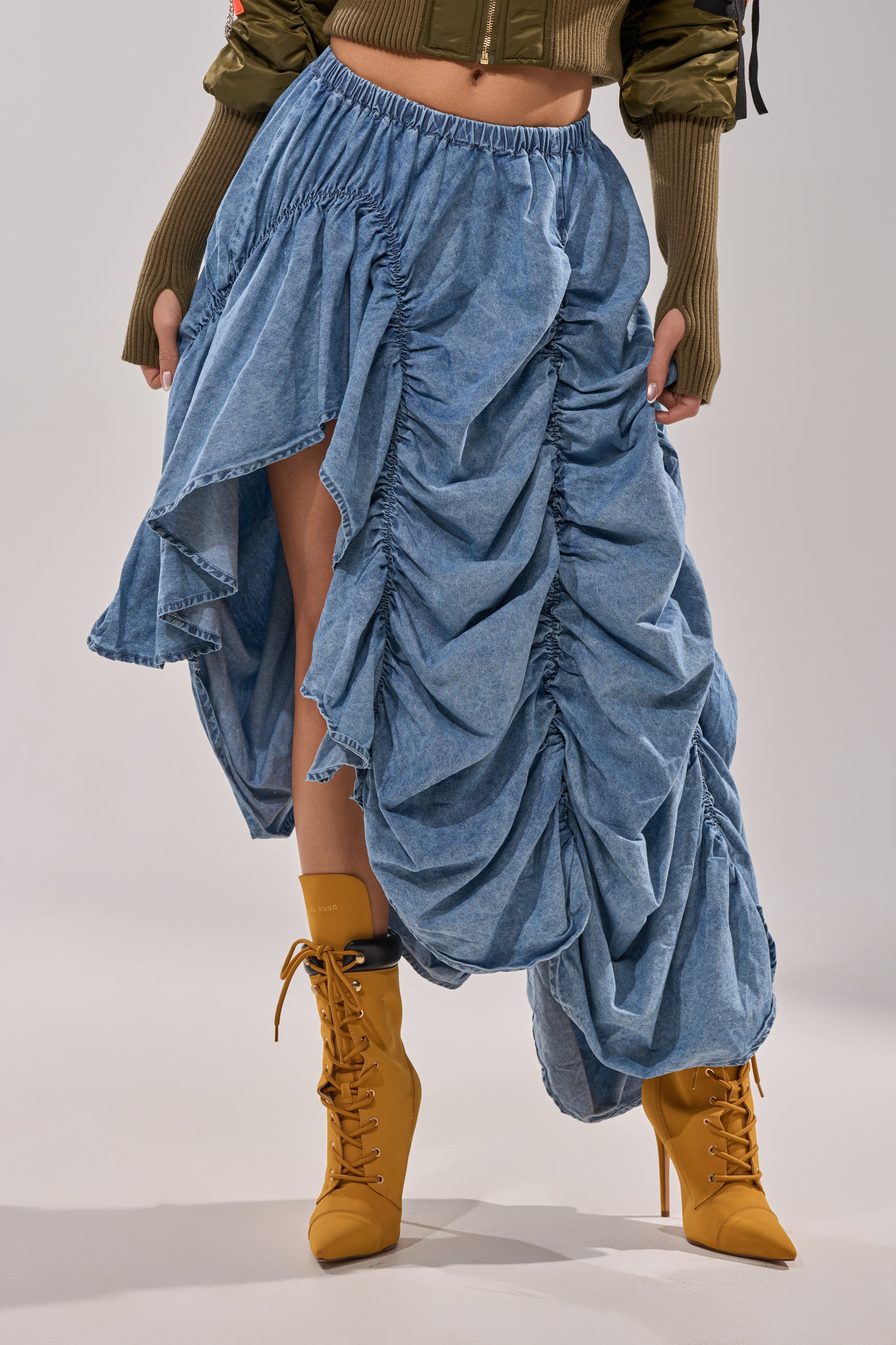 Someone wears the LETS CHANGE IT UP RUCHED MIDI SKIRT IN CHAMBRAY with mustard yellow lace-up boots and a green sweater, creating a modern, edgy look. 0
