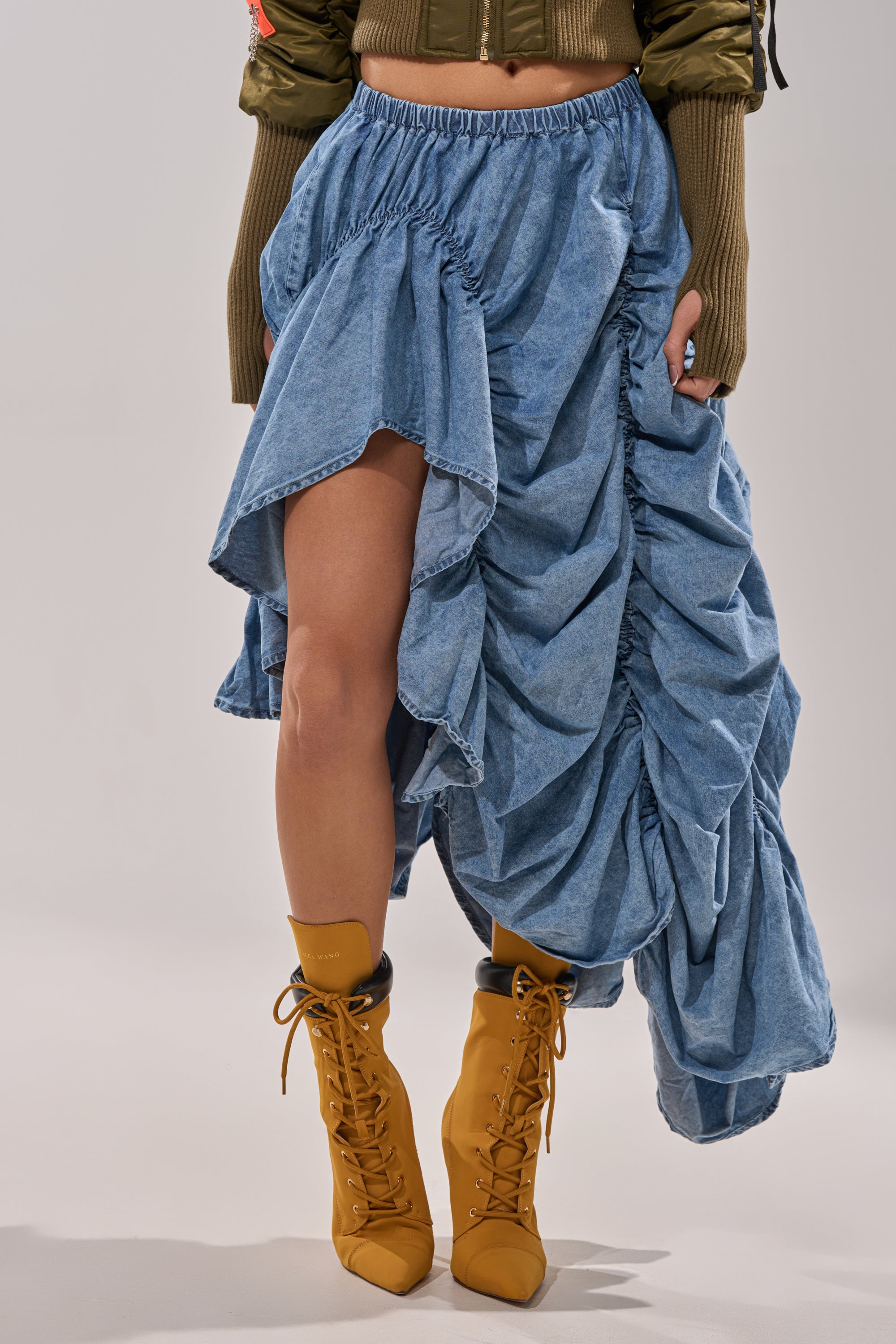 A person wears the LETS CHANGE IT UP RUCHED MIDI SKIRT IN CHAMBRAY, styled with mustard yellow heeled lace-up boots and a green long-sleeve cropped top, set against a plain light-colored background. 2
