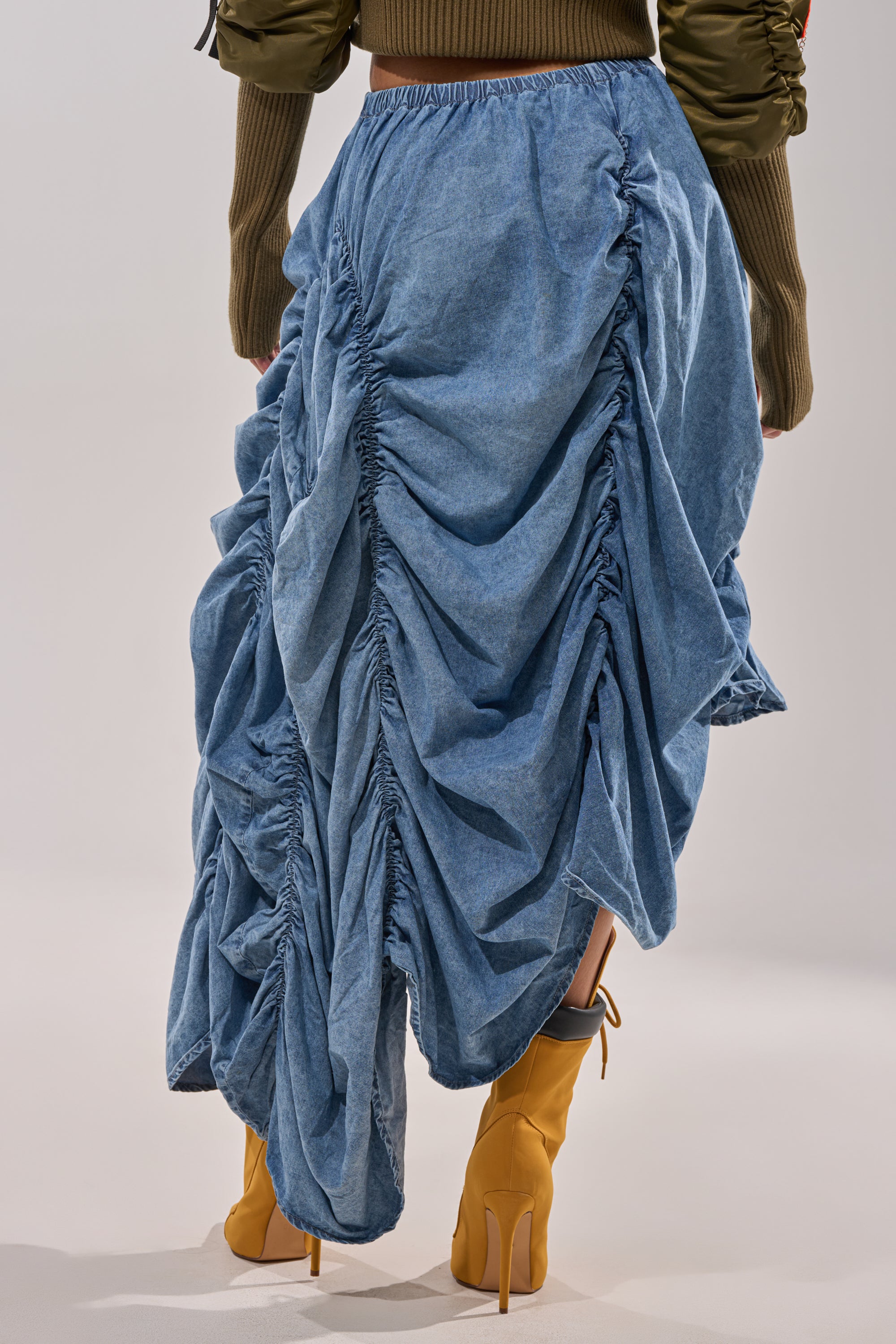 A person is shown from the back wearing the LETS CHANGE IT UP RUCHED MIDI SKIRT IN CHAMBRAY, styled with tan high-heeled boots and a long-sleeved olive green top. 5
