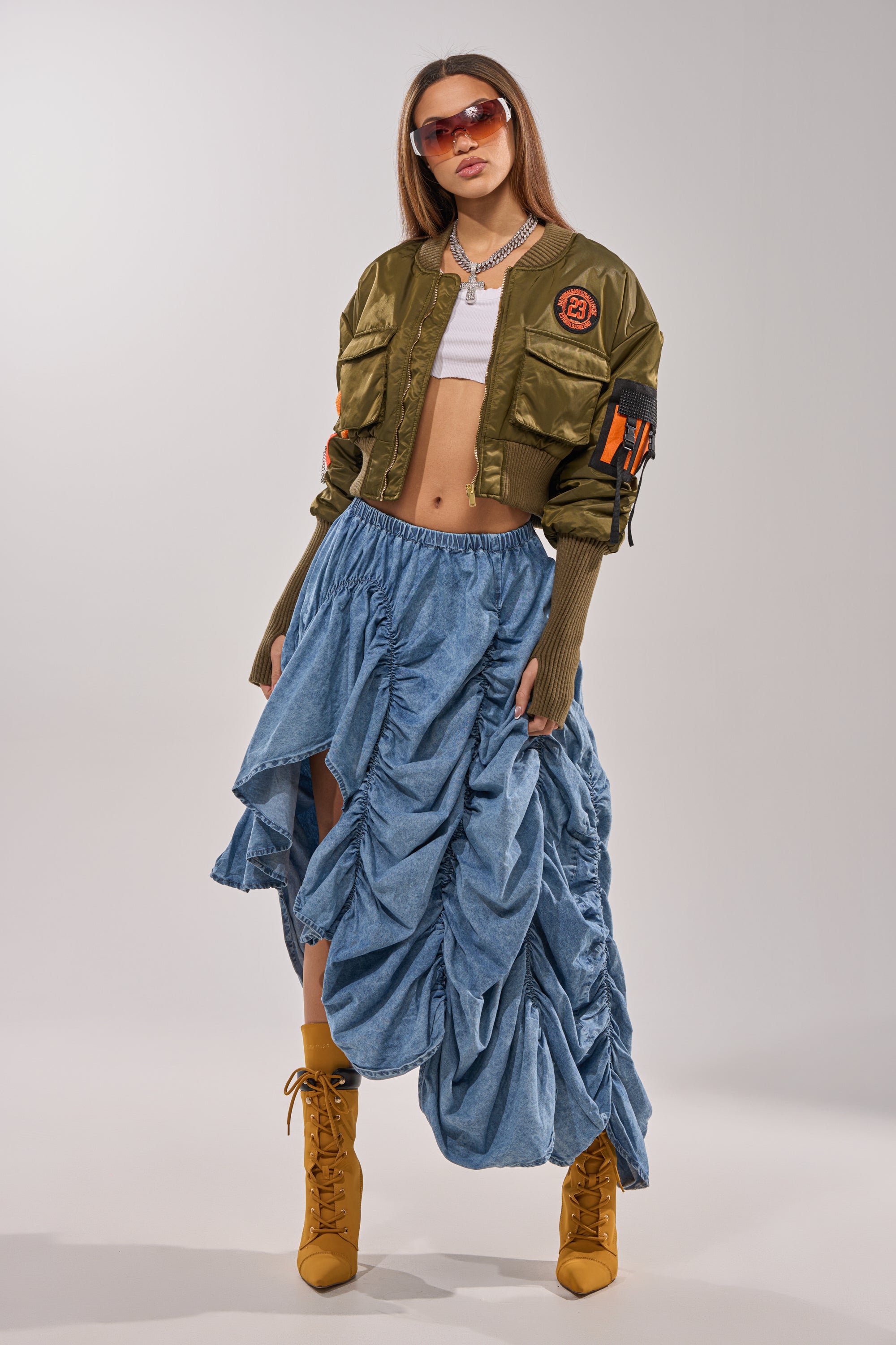 A woman poses in a cropped olive bomber jacket, white crop top, LETS CHANGE IT UP RUCHED MIDI SKIRT IN CHAMBRAY with high slits, tan lace-up boots, layered necklaces, and oversized sunglasses against a plain light background. 6
