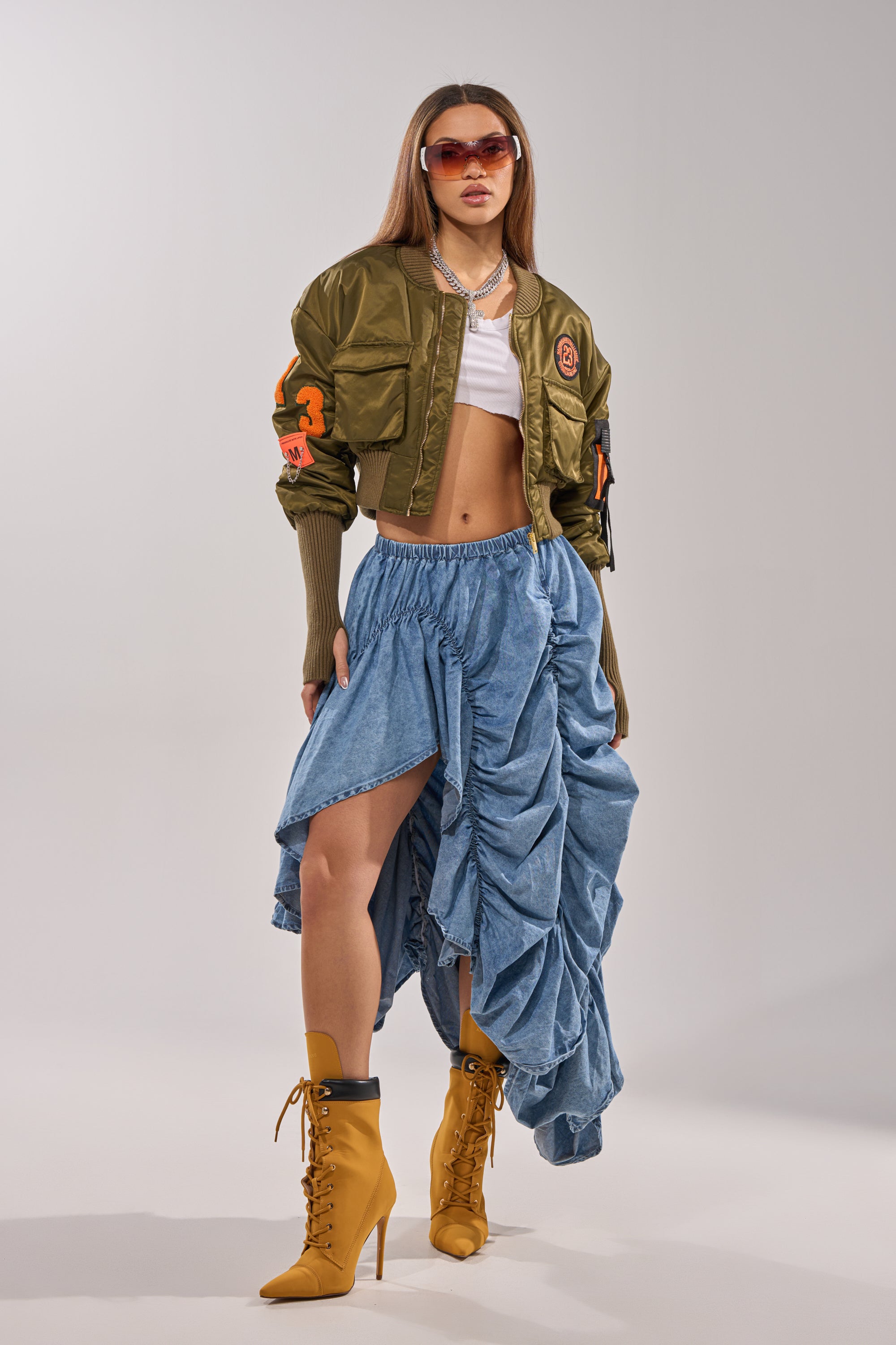 A woman wearing sunglasses, an olive green bomber jacket, a white crop top, the LETS CHANGE IT UP RUCHED MIDI SKIRT IN CHAMBRAY, and yellow lace-up high-heeled boots poses confidently against a simple light background. 7