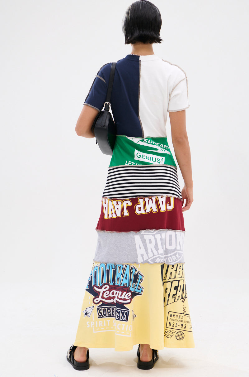 A person with short dark hair faces away, wearing the COOL GIRL ENERGY TSHIRT MAXI DRESS featuring colorful collegiate prints. They carry a black shoulder bag, wear black sandals, and stand against a plain white background.