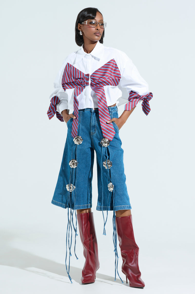 A person exudes confidence in GRUNGE ROSE DENIM BERMUDA SHORTS and a white shirt with oversized red-striped bow ties. Large circular decorations and tassels highlight their attire, paired with knee-high red boots. Sunglasses add flair to their short dark hair for a bold appearance.
