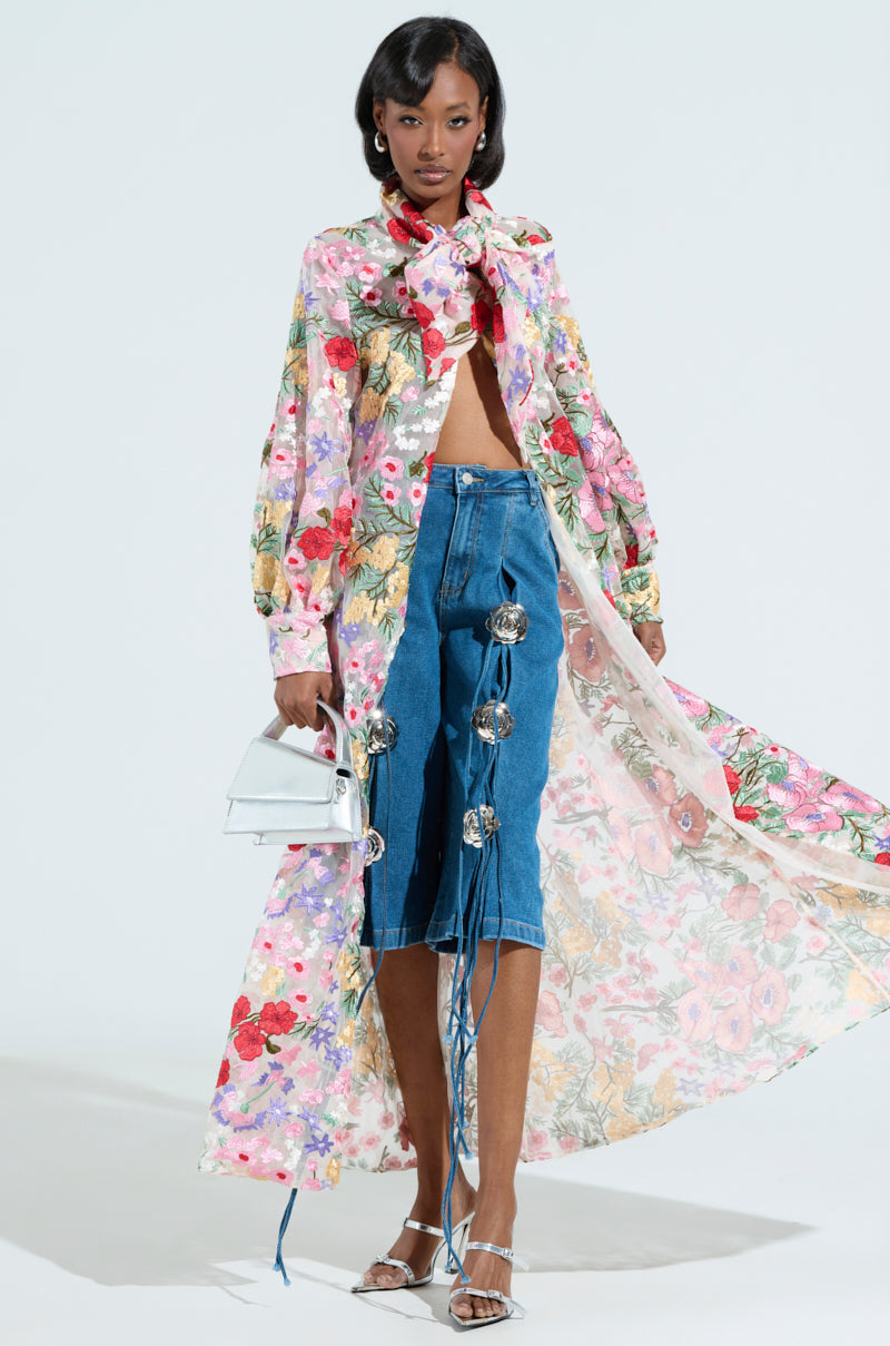 A person models a floral long-sleeve top paired with a matching scarf, showcasing Grunge Rose Denim Bermuda shorts decorated with large rhinestones. They accessorize with a silver handbag and silver heels while confidently posing against a plain backdrop.