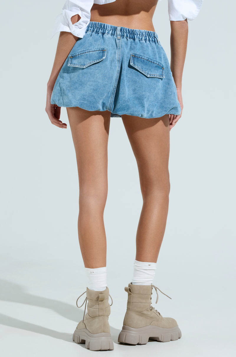 A person is wearing the ALL ABOUT IT denim bubble skirt in light blue, a white cropped top with cut-out details, tall white socks, and beige chunky lace-up boots. They are standing against a plain light background.