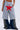 A person wears the SEE YOU NEXT TUESDAY PANT, flared plaid jeans with star-shaped metal studs at the hem, layered under athletic-inspired fleece shorts featuring contrast stripes, a red star patch, and a white drawstring. Hands rest in the shorts pockets.