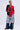 A person stands confidently in the SEE YOU NEXT TUESDAY PANT, paired with a split black and red jersey (#11), athletic fleece shorts with a red star, denim leg warmers with camo panels, hoop earrings, and a necklace against a plain background.