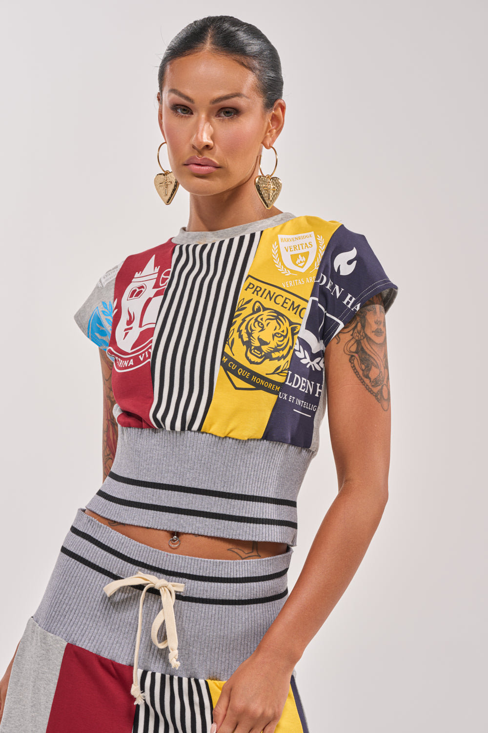 A woman with long dark hair in a ponytail models the MIXED FEELINGS CROPPED T-SHIRT, featuring vibrant patchwork, stripes, and crest designs. She pairs it with large heart-shaped earrings and stands confidently against a plain background. 0