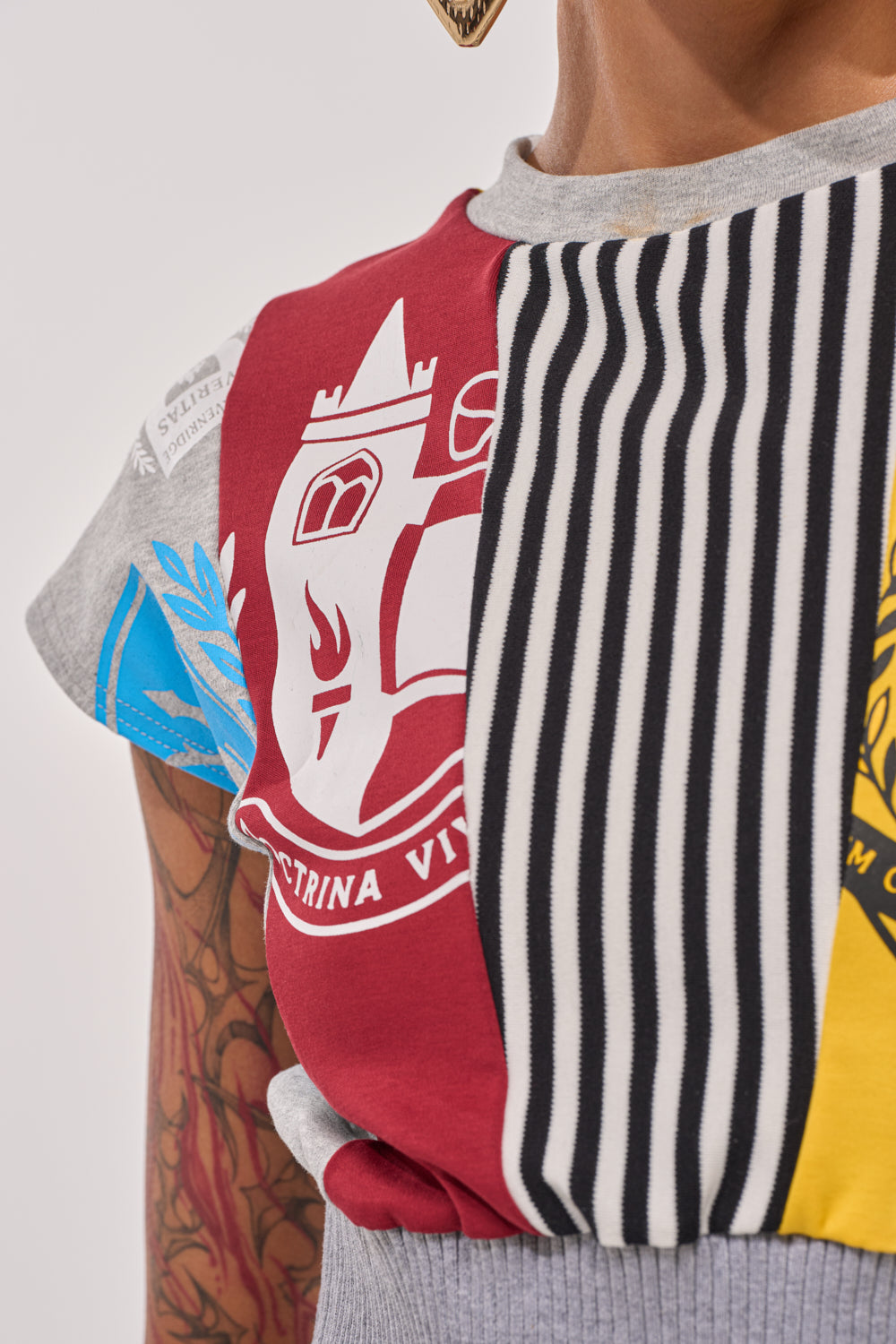 Close-up of a person wearing the MIXED FEELINGS CROPPED T-SHIRT—featuring vibrant patchwork: red with a white emblem, black and white stripes, blue and yellow accents. A tattooed arm adds to the bold streetwear vibe. 9