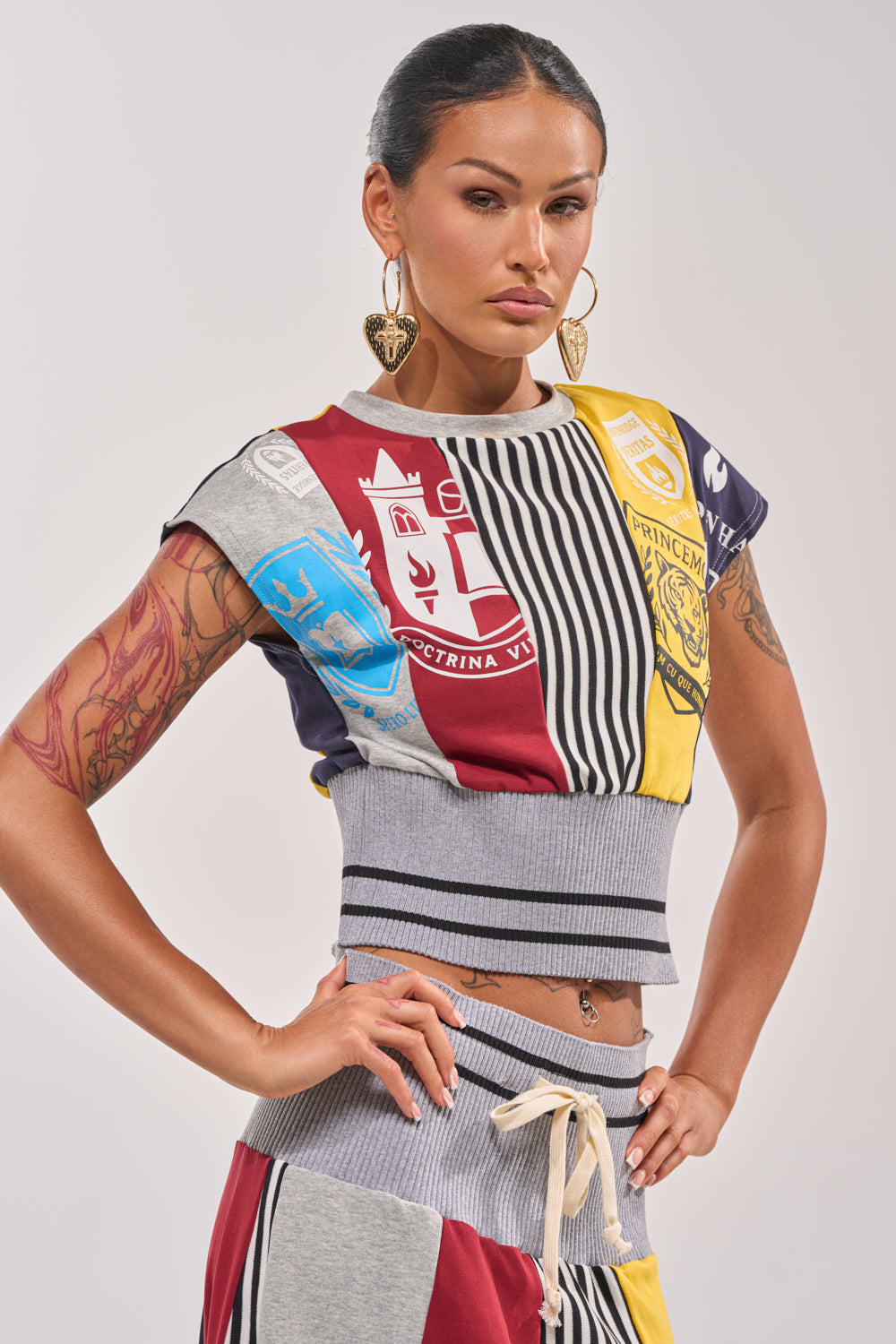 A tattooed woman wears the MIXED FEELINGS CROPPED T-SHIRT, featuring colorblock and patchwork patterns with crest designs. She has long dark hair tied back, gold heart-shaped earrings, and poses confidently against a plain background. 1