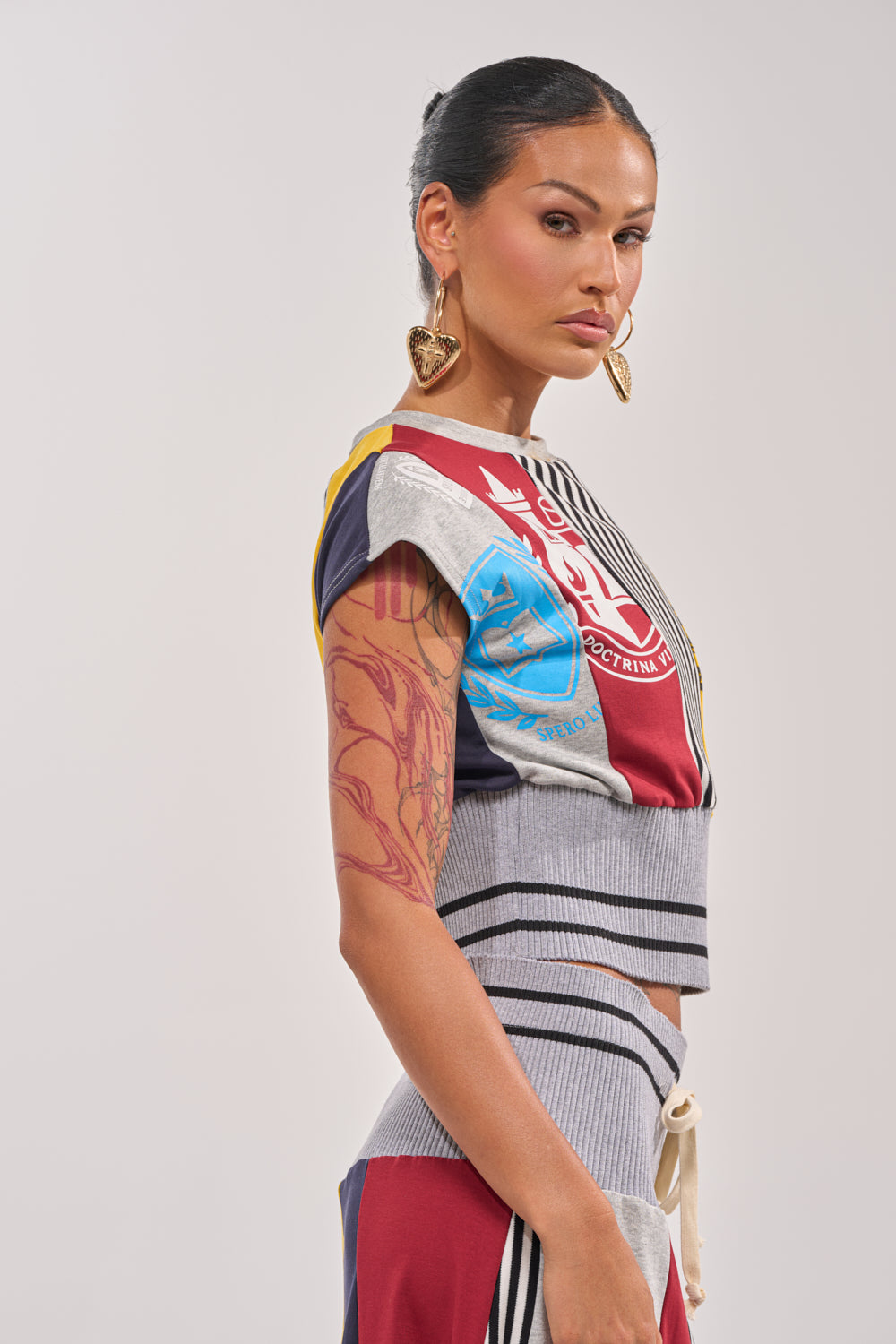 A woman with pulled-back dark hair, gold heart earrings, and a large upper arm tattoo stands sideways in the MIXED FEELINGS CROPPED T-SHIRT and matching pants with gray ribbed trim, looking confidently at the camera. 2