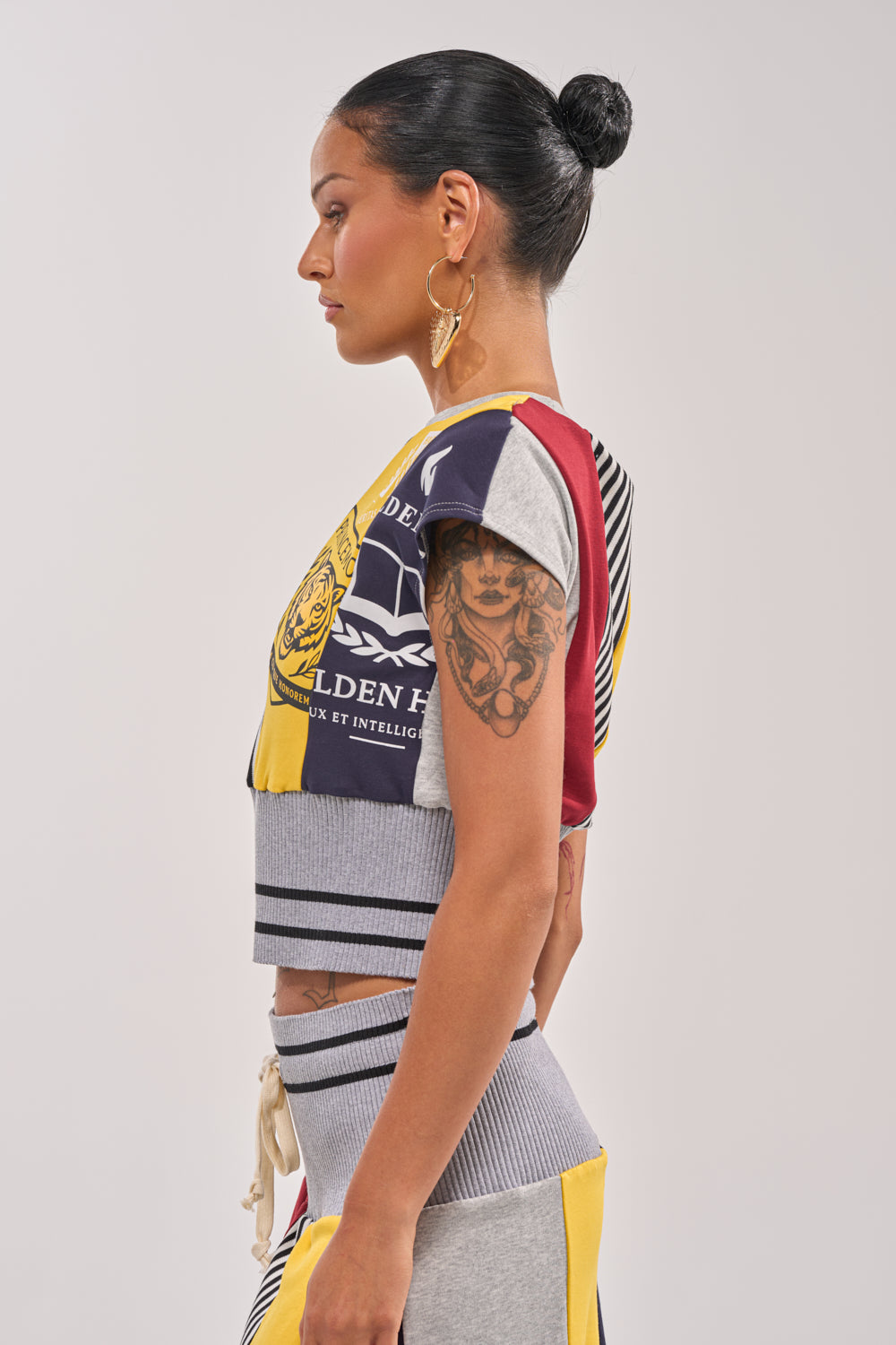 A woman with dark hair in a bun and gold hoop earrings stands in profile, wearing the MIXED FEELINGS CROPPED T-SHIRT and a skirt. She shows off a detailed upper arm tattoo against a neutral background. 3