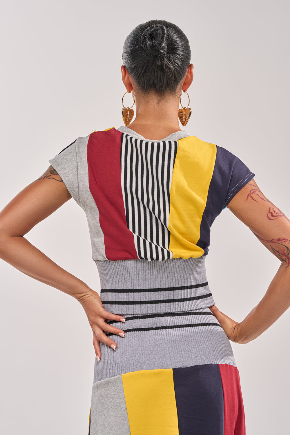 A woman with her hair in a neat bun and large heart-shaped earrings stands with her back to the camera, wearing the MIXED FEELINGS CROPPED T-SHIRT—a bold colorblock varsity top with gray, yellow, red, black, and white stripes. 4