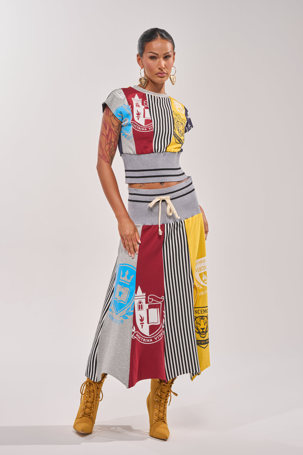 A woman stands confidently in the MIXED FEELINGS CROPPED T-SHIRT and matching skirt with colorful Hogwarts house crests, vertical stripes, and patchwork patterns. She pairs it with tan boots, hoop earrings, and a tied-back hairstyle against a light gray background. 7