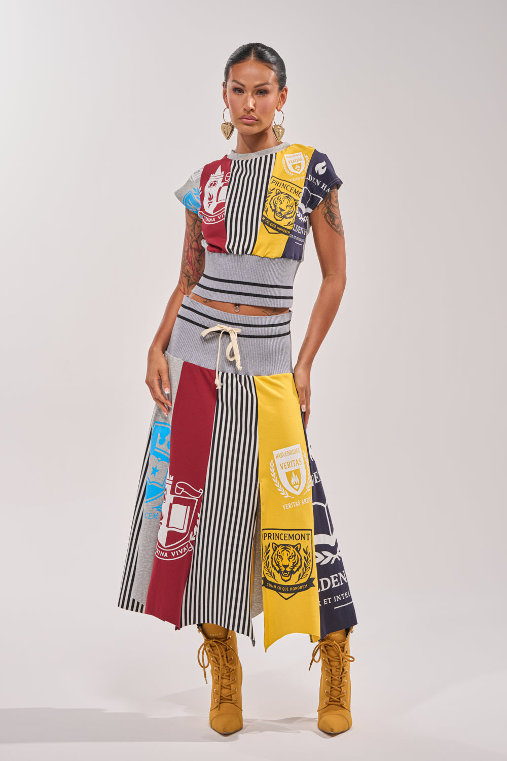 A woman confidently wears the MIXED FEELINGS MIDI SKIRT—a colorful, sporty patchwork piece with crests, stripes, and text—paired with a sleeveless top, yellow lace-up boots, and large hoop earrings against a plain background.