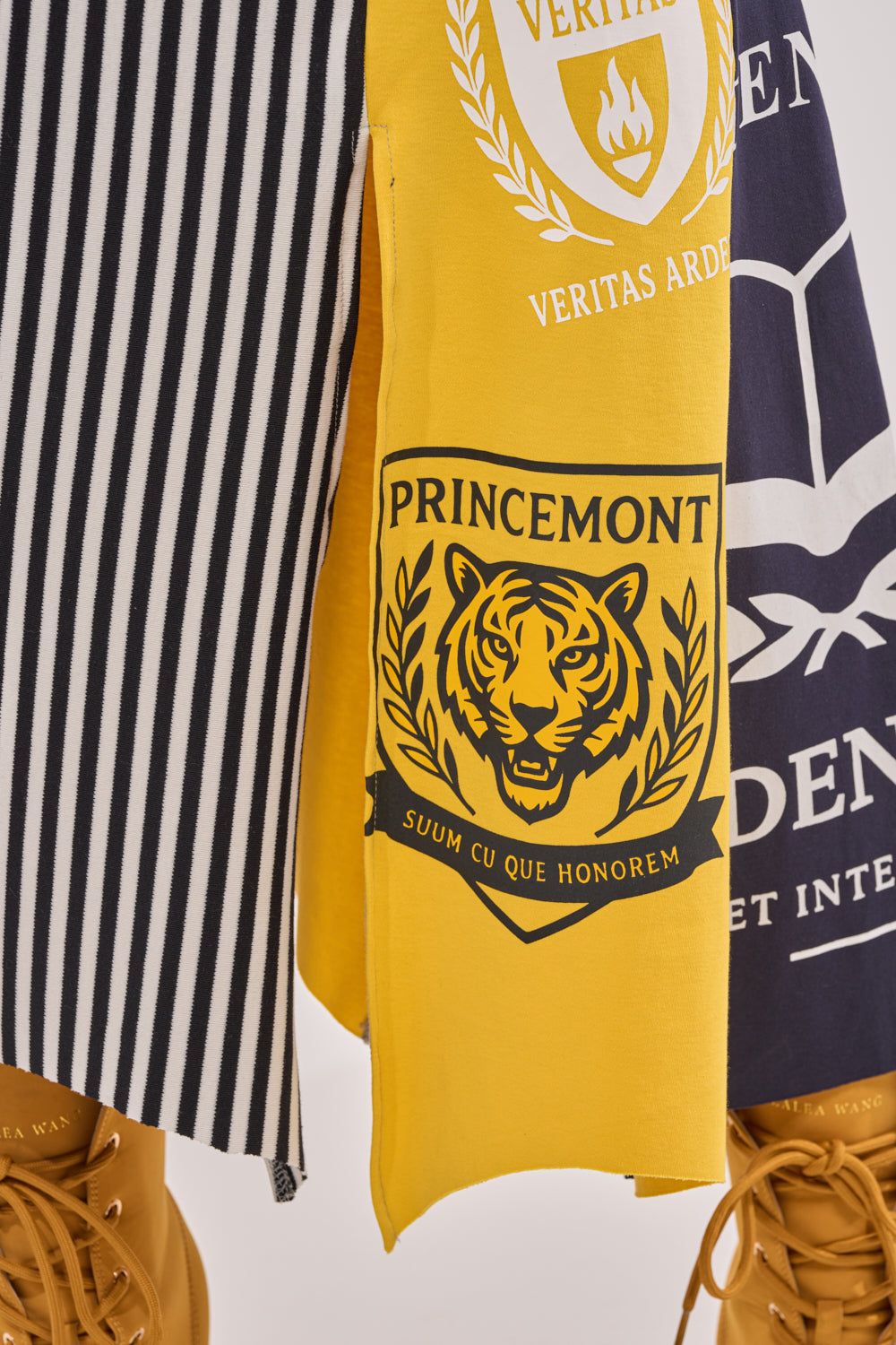 Close-up of two skirts: the MIXED FEELINGS MIDI SKIRT featuring black and white vertical patchwork stripes, and a yellow colorblock varsity skirt with tiger emblem, laurel leaves, and "PRINCEMONT - SUUM CU QUE HONOREM"; paired with brown lace-up boots. 9