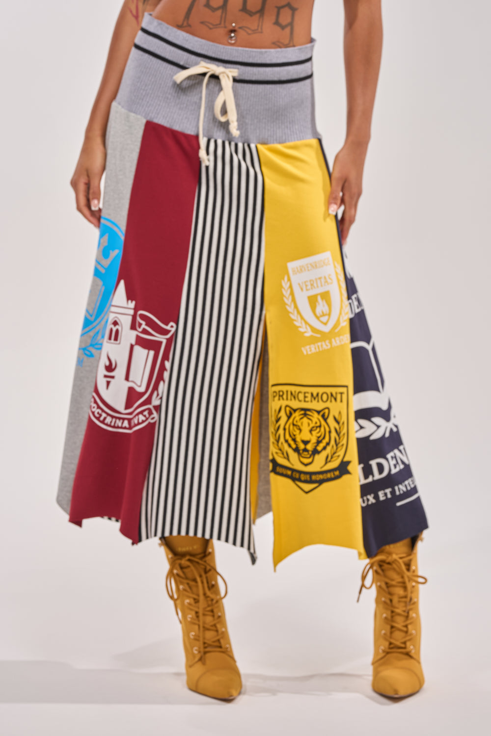 Wearing the MIXED FEELINGS MIDI SKIRT featuring bold colorblock stripes, crests, and a drawstring waist, paired with yellow lace-up boots and showing a tattooed torso, this look channels sporty streetwear energy.