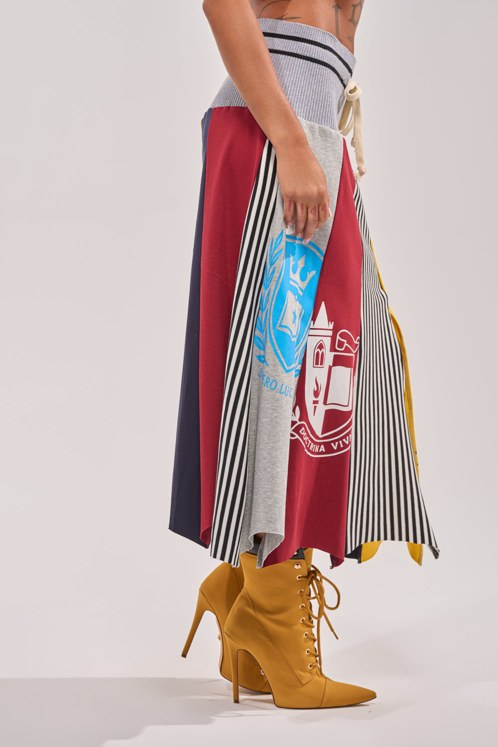 Only the lower half of a person in profile is shown wearing the MIXED FEELINGS MIDI SKIRT, featuring colorful patchwork and emblems, paired with pointed yellow high-heeled boots.
