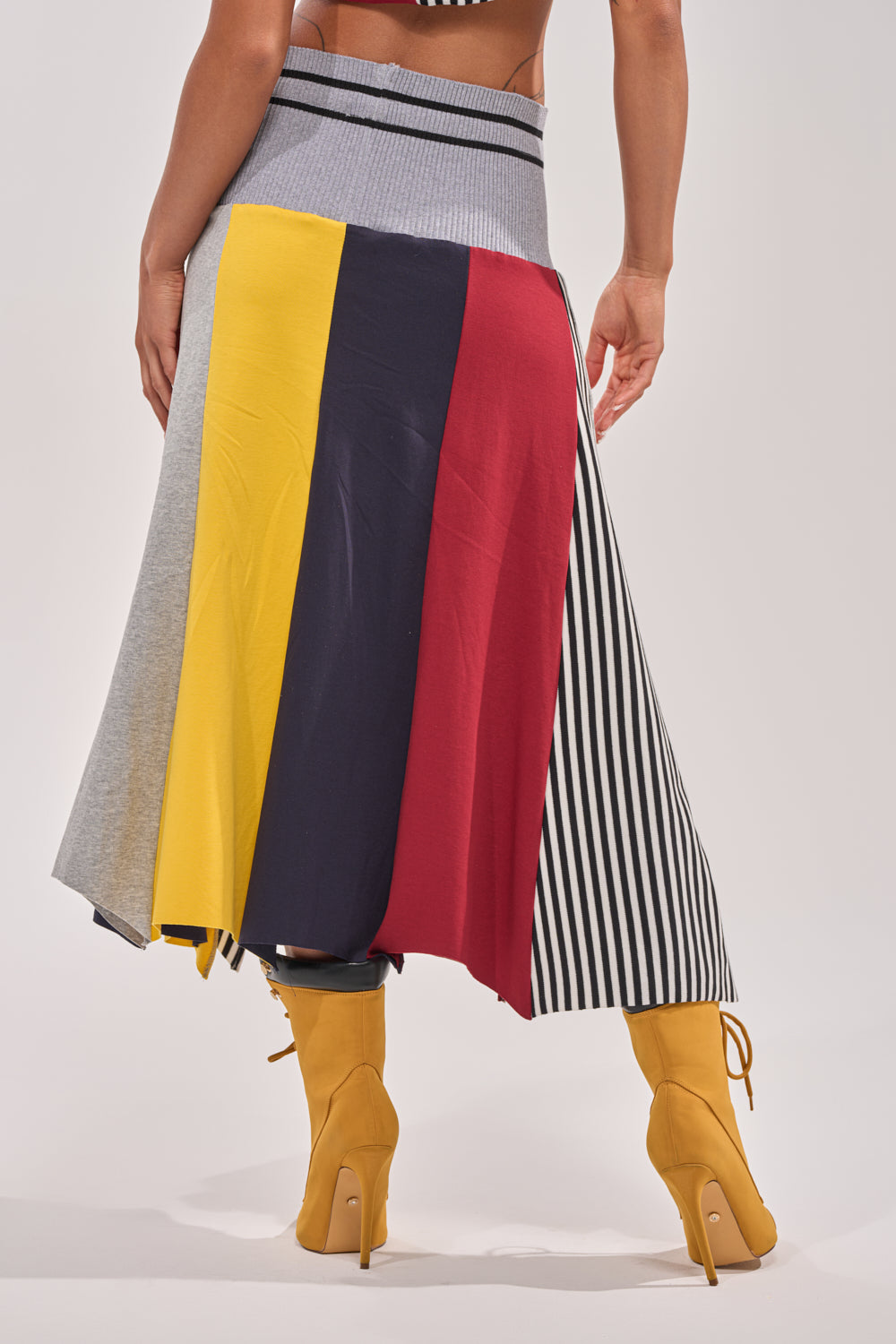 A person rocking the MIXED FEELINGS MIDI SKIRT with gray, yellow, navy, red, and black-and-white vertical panels and pairing it with mustard yellow heeled boots for a sporty streetwear look. 5