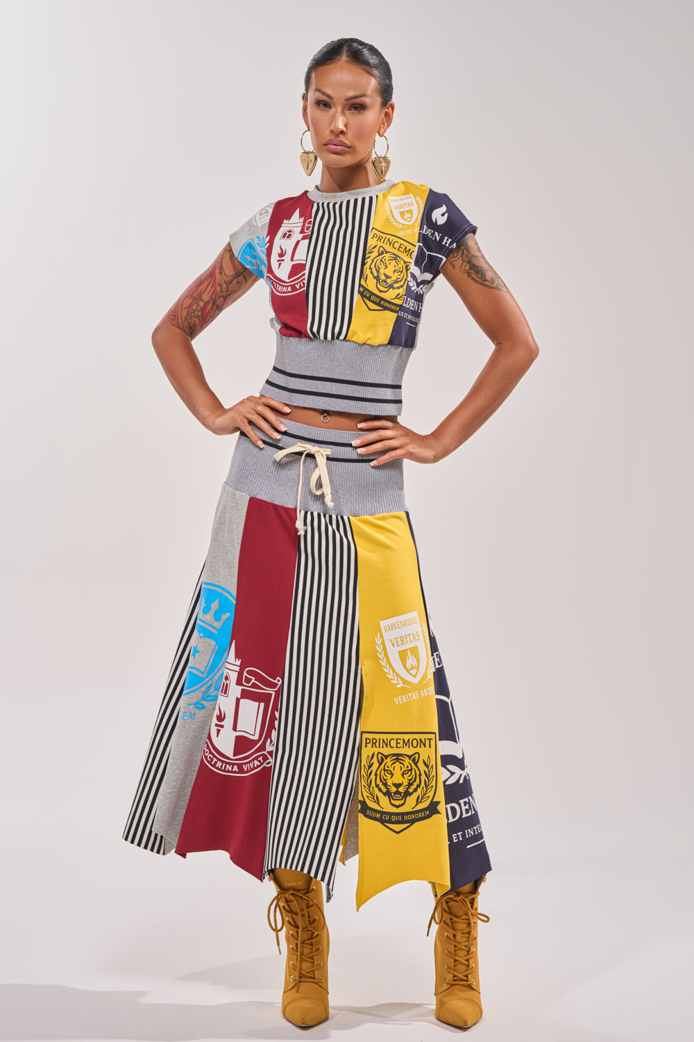 A woman faces forward in the MIXED FEELINGS MIDI SKIRT, featuring colorful varsity patchwork with crest and stripe patterns. She pairs it with a cropped top, tan lace-up boots, hoop earrings, and displays arm tattoos for a sporty streetwear vibe. 6
