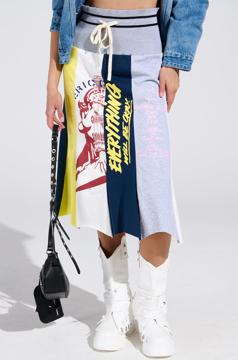 Wearing the REBUILT MIDI SKIRT with bold graphics, paired with a denim jacket, white boots, and holding a small black handbag; only the lower torso and legs are visible.
