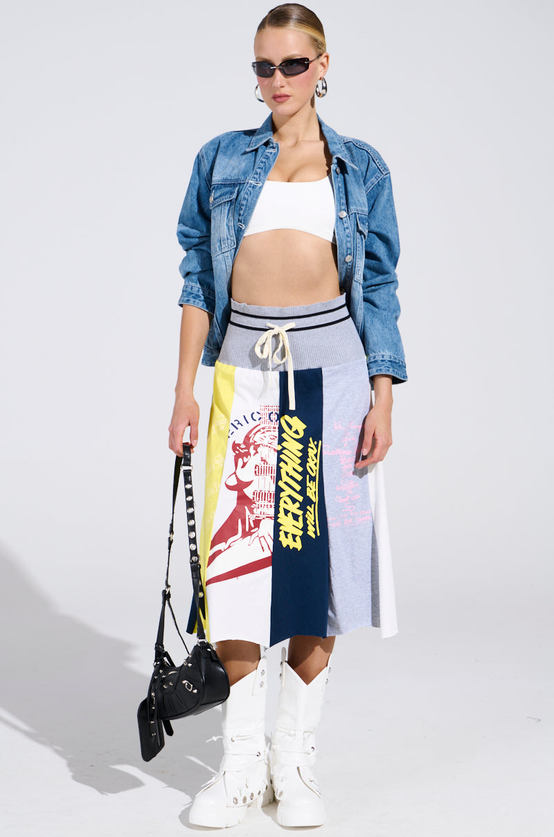 A woman in sunglasses wears a denim jacket, white bandeau top, and the REBUILT MIDI SKIRT featuring text and graphics. She completes her sporty-chic look with white boots, hoop earrings, and a black handbag.