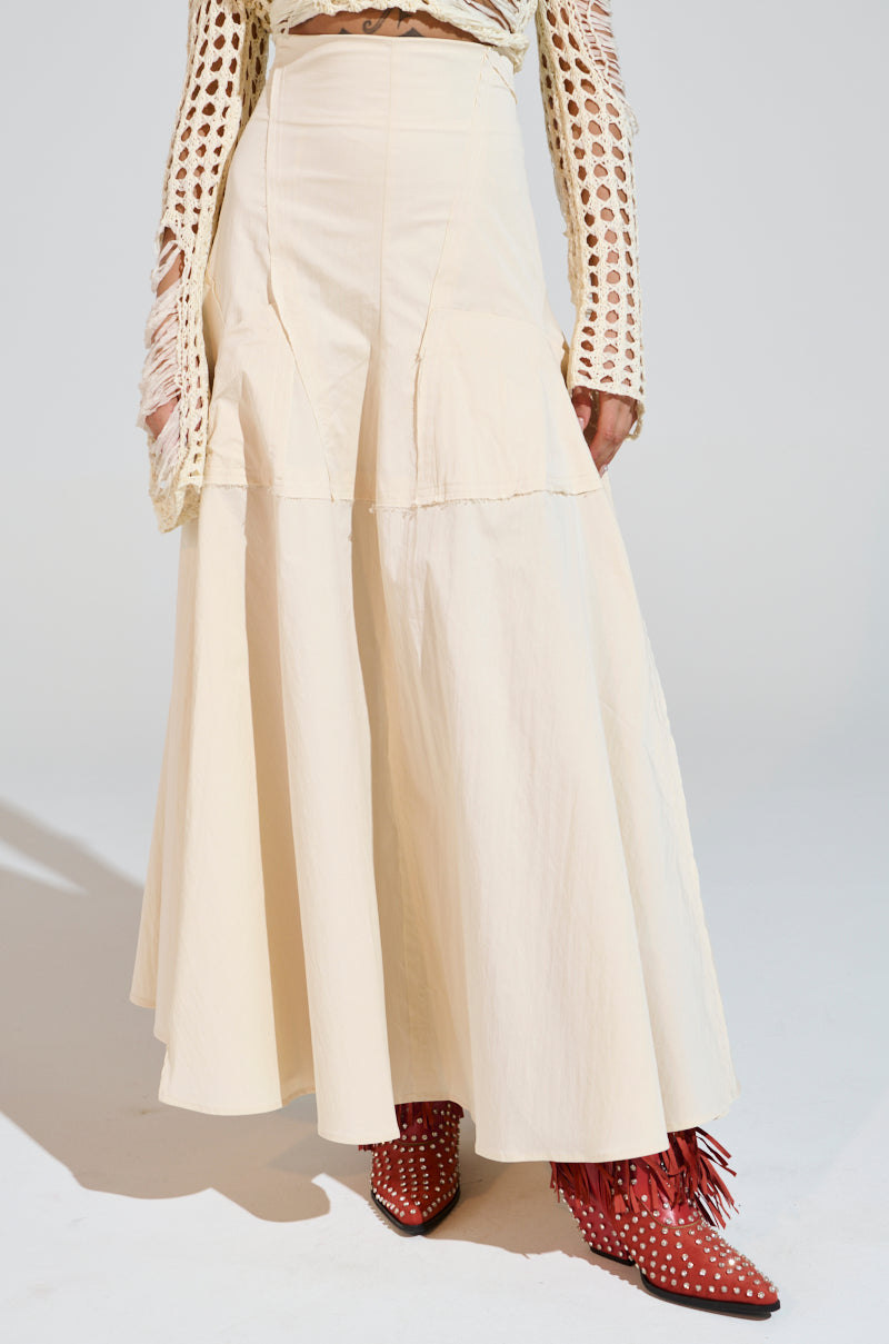 The individual is wearing a CLAIRE EXPOSED SEAMS MIDI SKIRT, a cream-colored A-line with a high waist, and a textured beige top with cutouts. They stylishly complement it with red studded fringe boots on a plain light gray background. 2