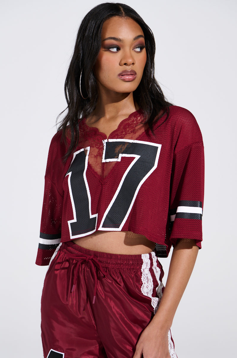 Sporting a LET'S GET IT JERSEY in maroon with the number 17, coupled with matching shorts adorned with white lace on the sides, they present a dynamic look in a cropped oversized silhouette, posing thoughtfully against a plain backdrop.