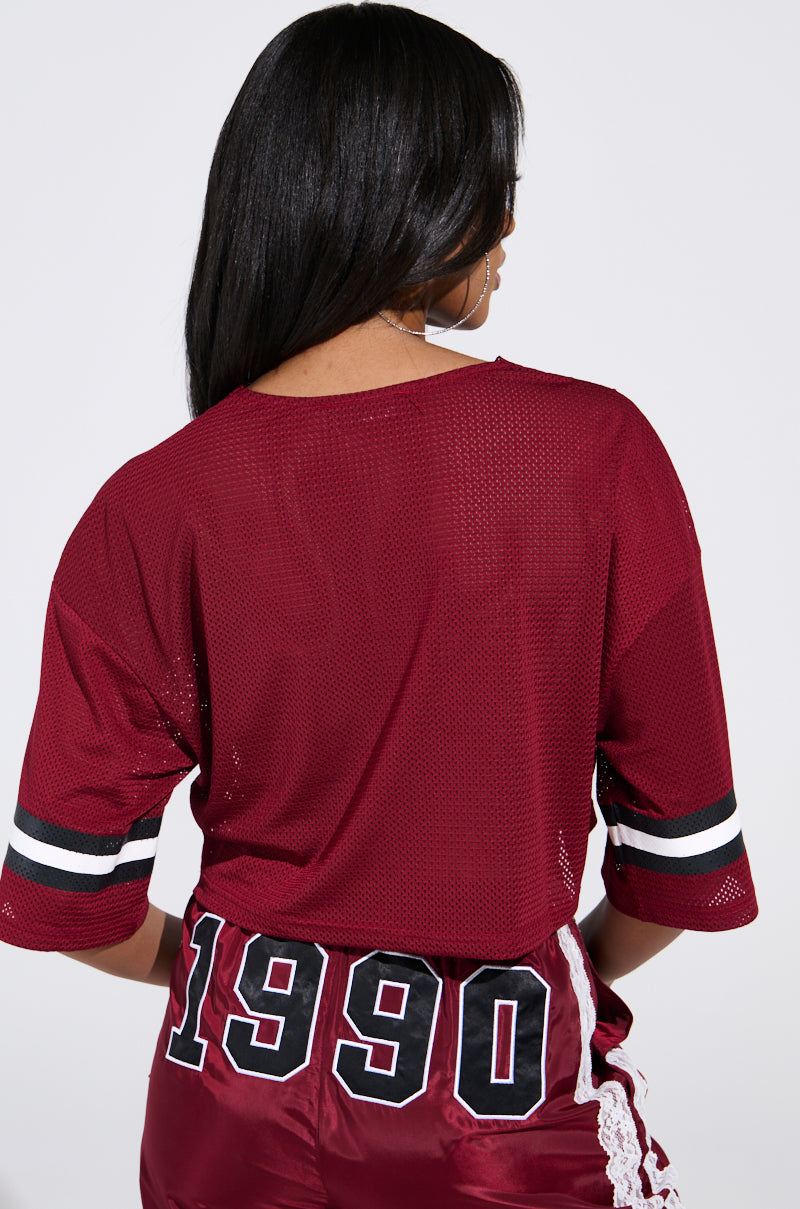A person with long hair faces away, wearing the LETS GET IT JERSEY, a maroon mesh top with black and white striped sleeves. Their sporty-chic maroon shorts showcase the number "1990.